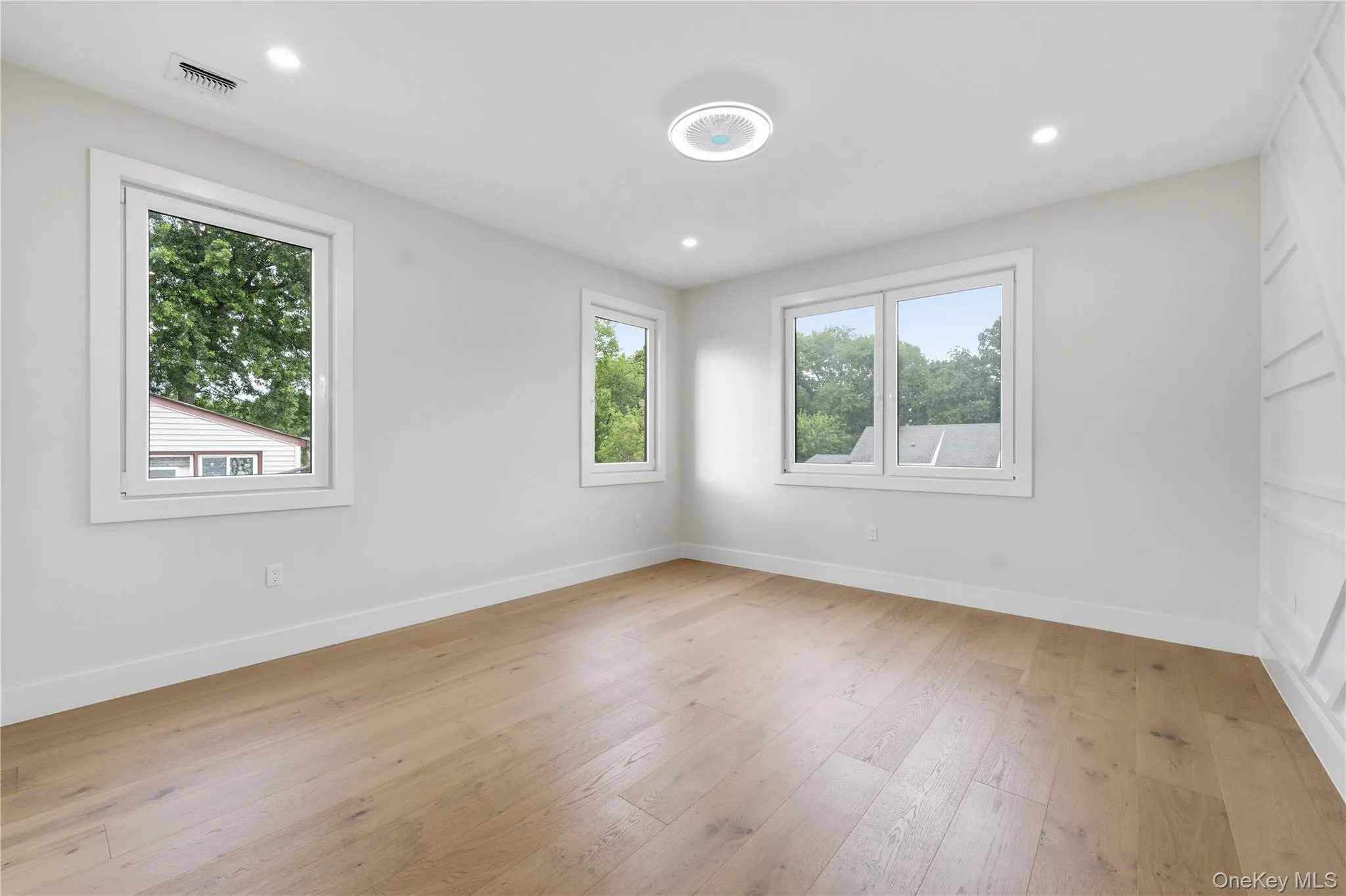 Bright room featuring light wood-finish flooring, crisp white walls, three windows with white trim, recessed ceiling lighting, and a modern flush-mount ceiling fixture and custom mill work wainscoting wall Bright room featuring light wood-finish flooring, crisp white walls, three windows with white trim, recessed ceiling lighting, and a modern flush-mount ceiling fixture and custom mill work wainscoting wall