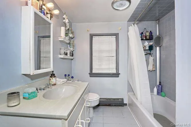 Full bath with shower / bath combination with curtain, vanity, toilet, a baseboard radiator, and tile patterned floors Full bath with shower / bath combination with curtain, vanity, toilet, a baseboard radiator, and tile patterned floors