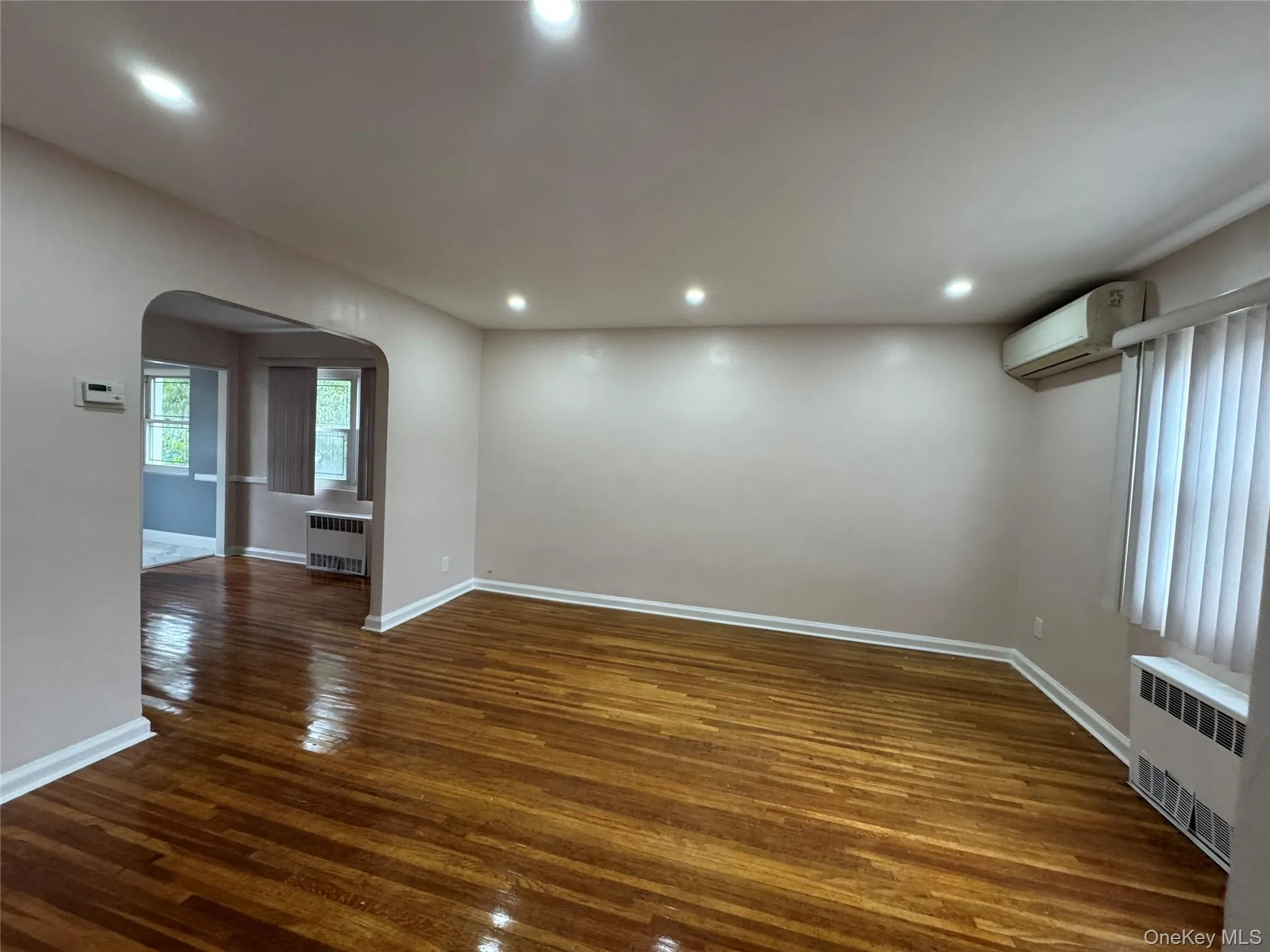Empty room with arched walkways, dark wood-style flooring, radiator heating unit, recessed lighting, and an AC wall unit Empty room with arched walkways, dark wood-style flooring, radiator heating unit, recessed lighting, and an AC wall unit