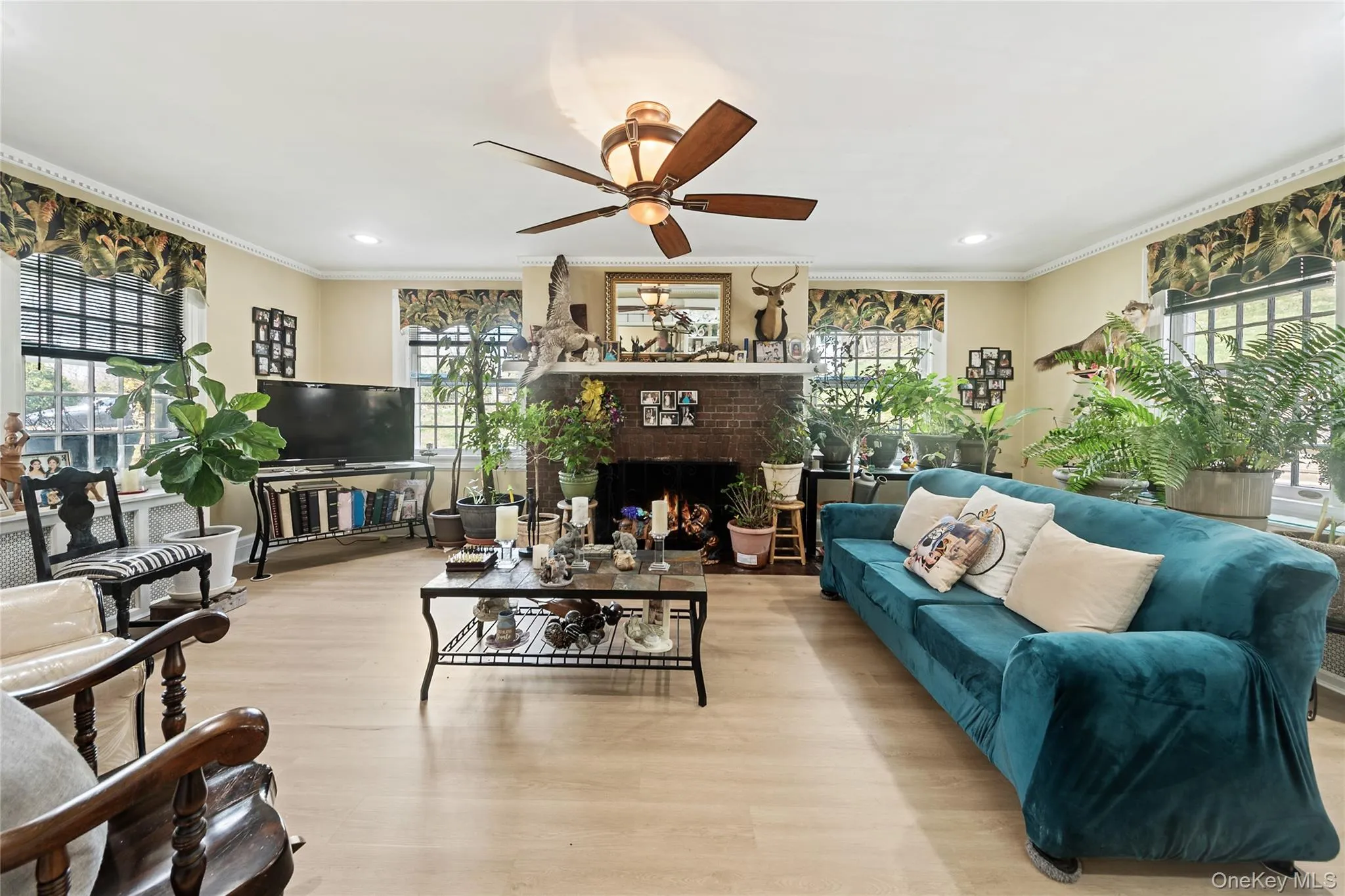 Spacious living area featuring a brick fireplace, light wood-finish flooring, crown molding, a ceiling fan, and recessed lighting Spacious living area featuring a brick fireplace, light wood-finish flooring, crown molding, a ceiling fan, and recessed lighting
