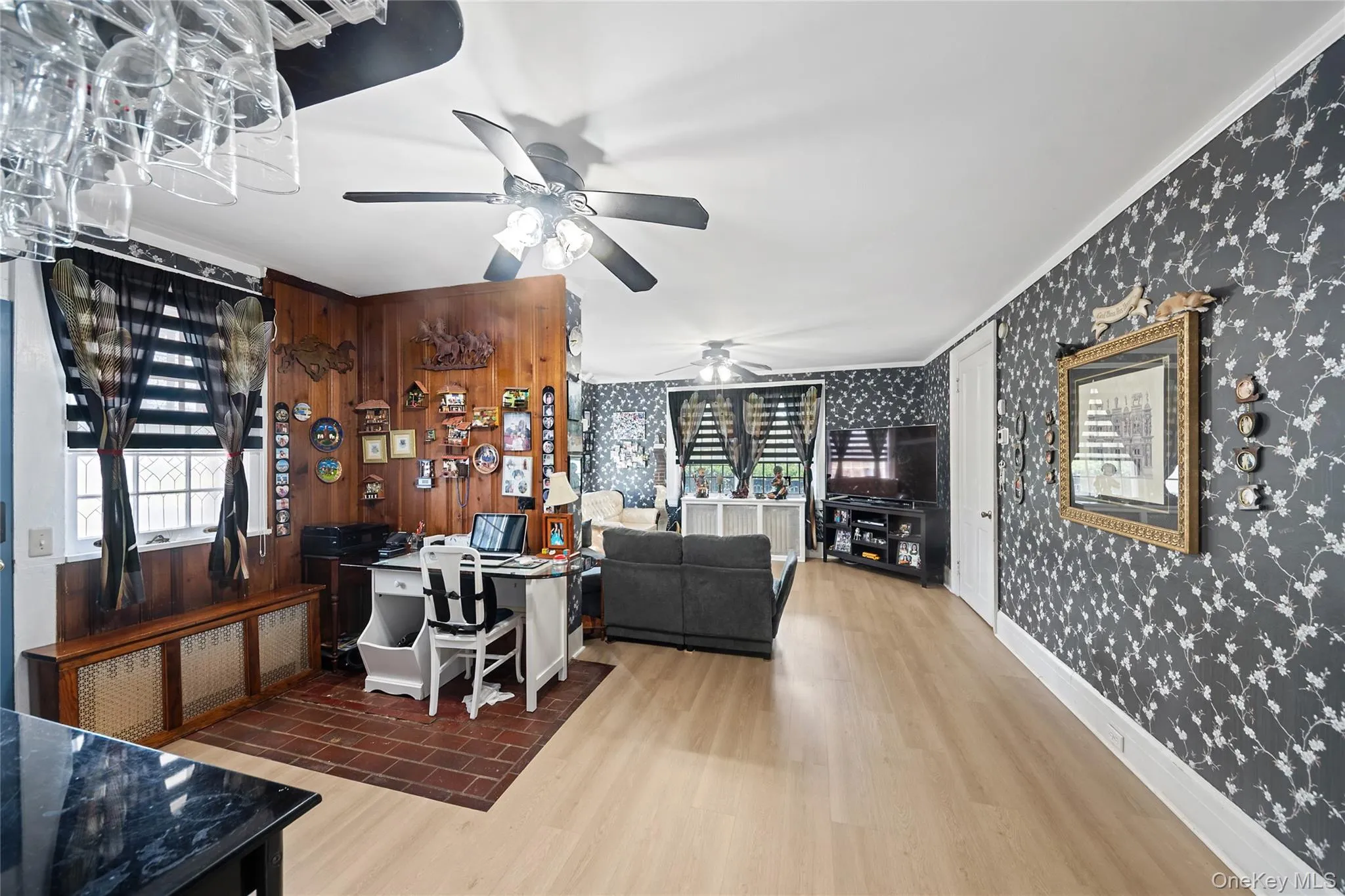 Open-concept living area featuring light wood-finish flooring, a ceiling fan with integrated lighting, and dark floral wallpaper Open-concept living area featuring light wood-finish flooring, a ceiling fan with integrated lighting, and dark floral wallpaper