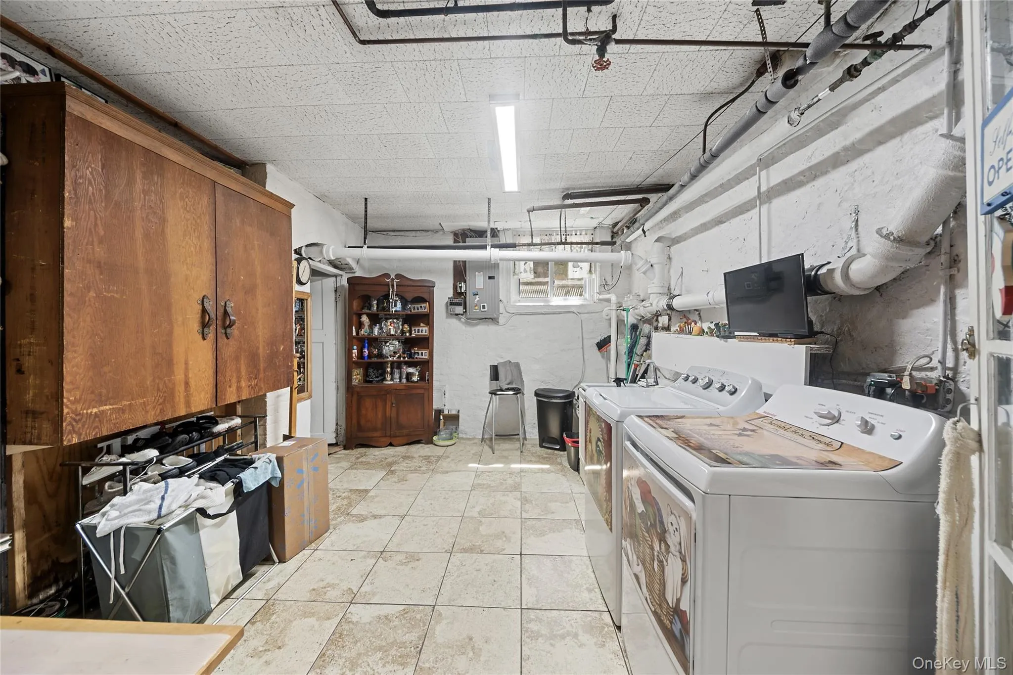 Utility space featuring tiled flooring, exposed ceiling pipes, white painted brick walls, two laundry appliances, and built-in wood cabinetry Utility space featuring tiled flooring, exposed ceiling pipes, white painted brick walls, two laundry appliances, and built-in wood cabinetry