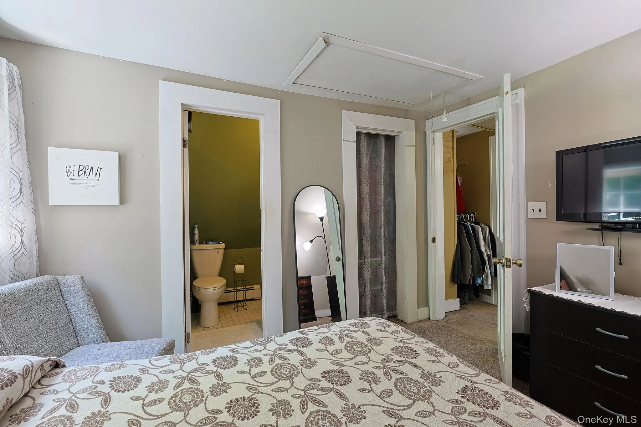 Bedroom featuring a window with sheer curtains, a built-in closet with carpet flooring, and an en-suite bathroom with a toilet and white tile flooring Bedroom featuring a window with sheer curtains, a built-in closet with carpet flooring, and an en-suite bathroom with a toilet and white tile flooring