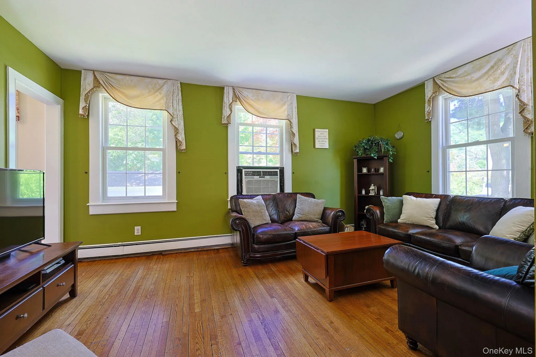 Wood-finish flooring throughout, featuring multiple double-hung windows with white trim, baseboard heating, and a built-in shelf unit Wood-finish flooring throughout, featuring multiple double-hung windows with white trim, baseboard heating, and a built-in shelf unit
