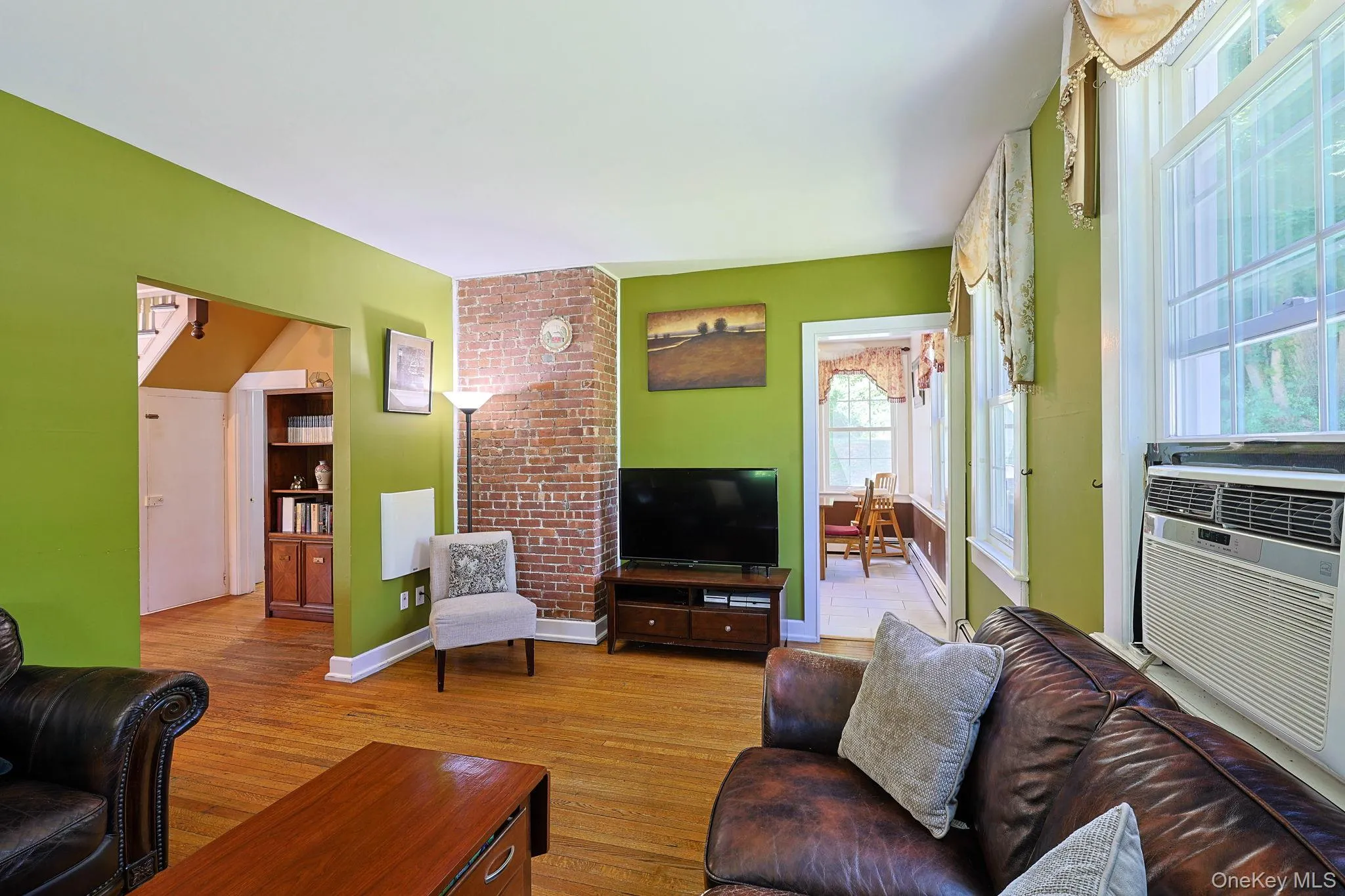 Living area featuring wood-finish flooring, exposed brick accent wall, multiple windows, and a built-in window air conditioning unit Living area featuring wood-finish flooring, exposed brick accent wall, multiple windows, and a built-in window air conditioning unit