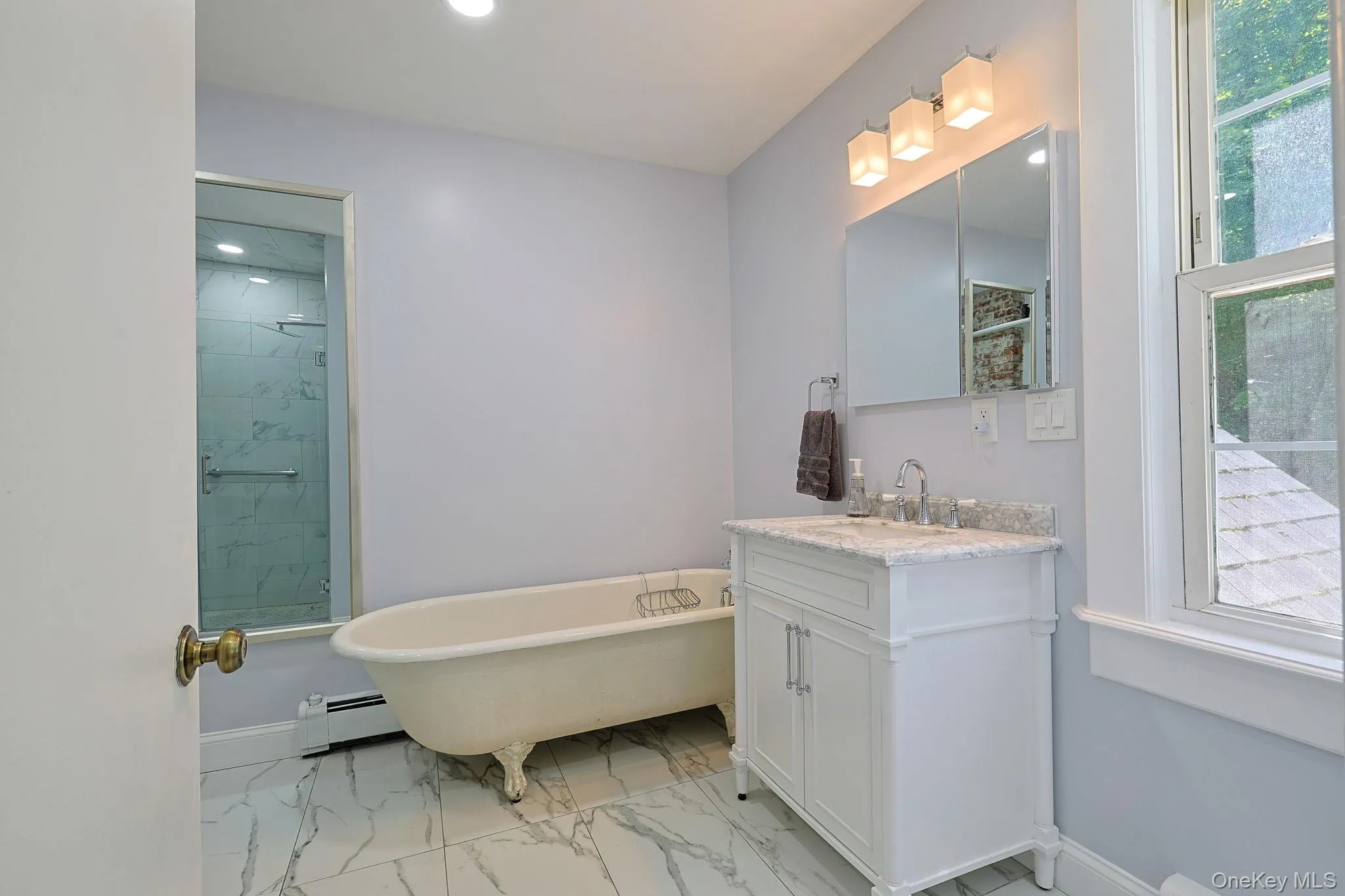 Bathroom featuring a freestanding clawfoot tub, a white vanity with a light-toned countertop, a mirrored medicine cabinet, a glass-enclosed shower with marble-look tile, and white marble-look floor tile Bathroom featuring a freestanding clawfoot tub, a white vanity with a light-toned countertop, a mirrored medicine cabinet, a glass-enclosed shower with marble-look tile, and white marble-look floor tile