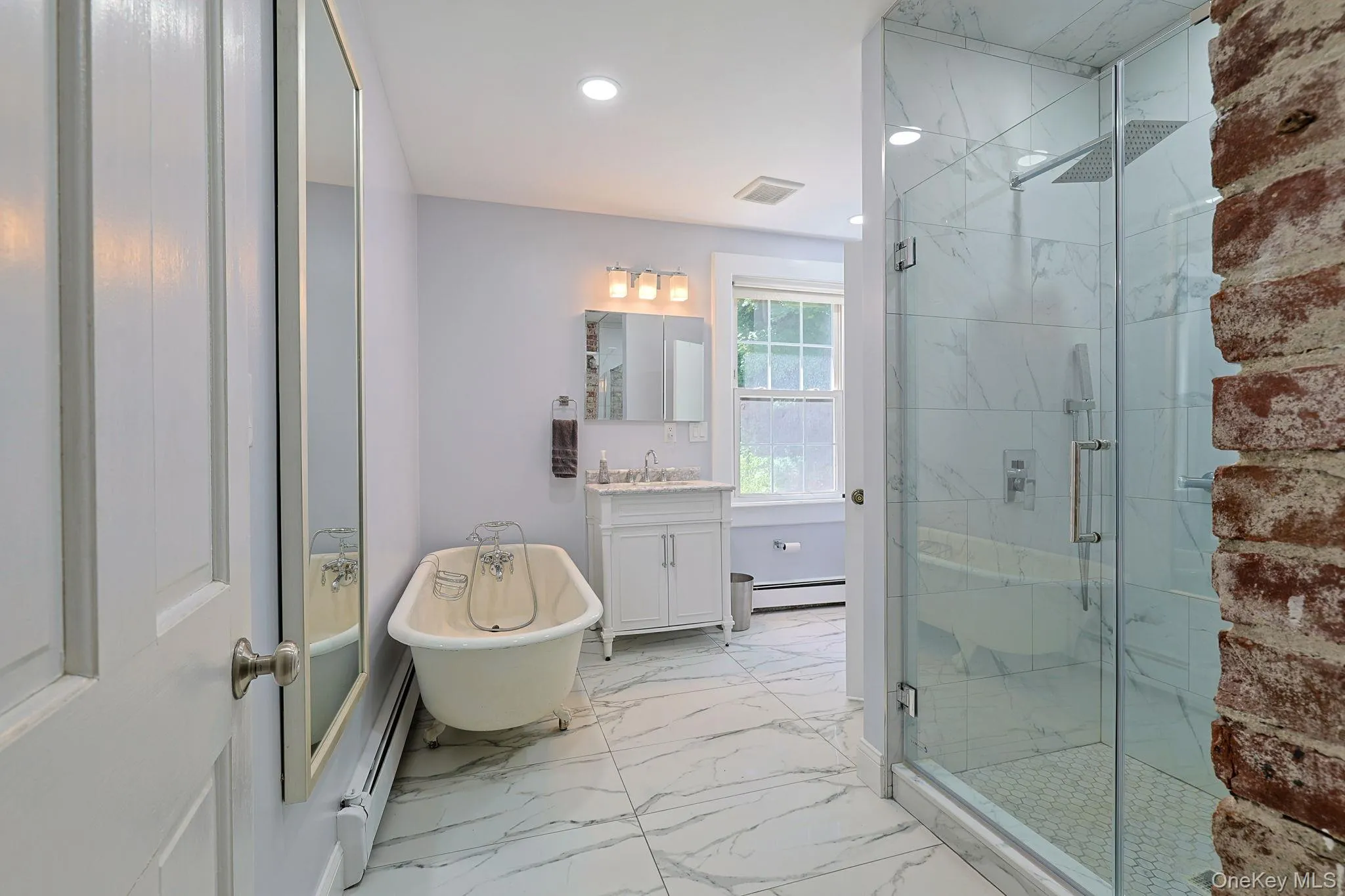 Bathroom featuring a glass-enclosed shower with rainfall showerhead, white marble-style tile, freestanding clawfoot bathtub, and a single vanity with a white countertop Bathroom featuring a glass-enclosed shower with rainfall showerhead, white marble-style tile, freestanding clawfoot bathtub, and a single vanity with a white countertop