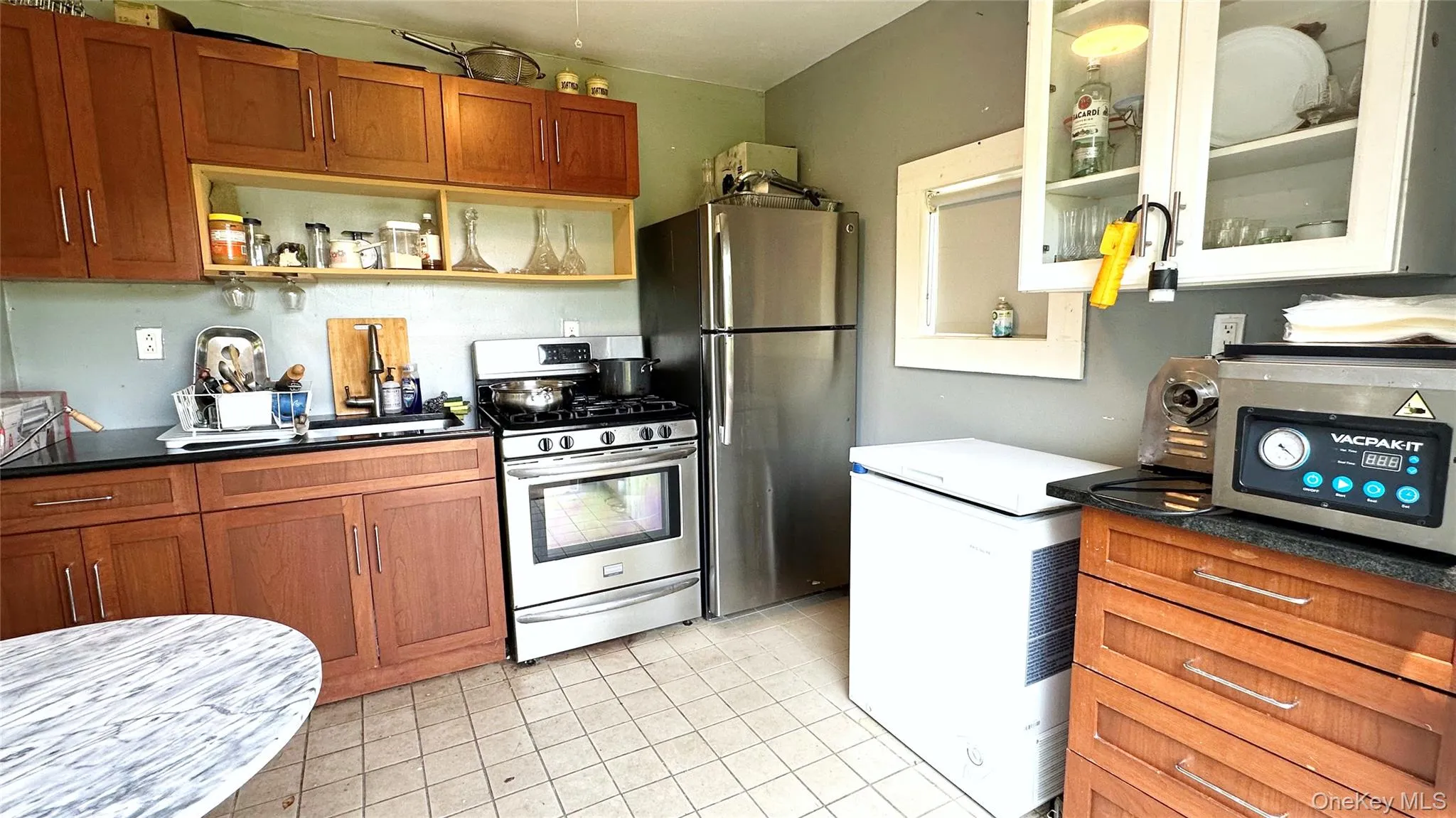 Kitchen featuring appliances with stainless steel finishes, brown cabinets, glass insert cabinets, light tile patterned floors, and open shelves Kitchen featuring appliances with stainless steel finishes, brown cabinets, glass insert cabinets, light tile patterned floors, and open shelves
