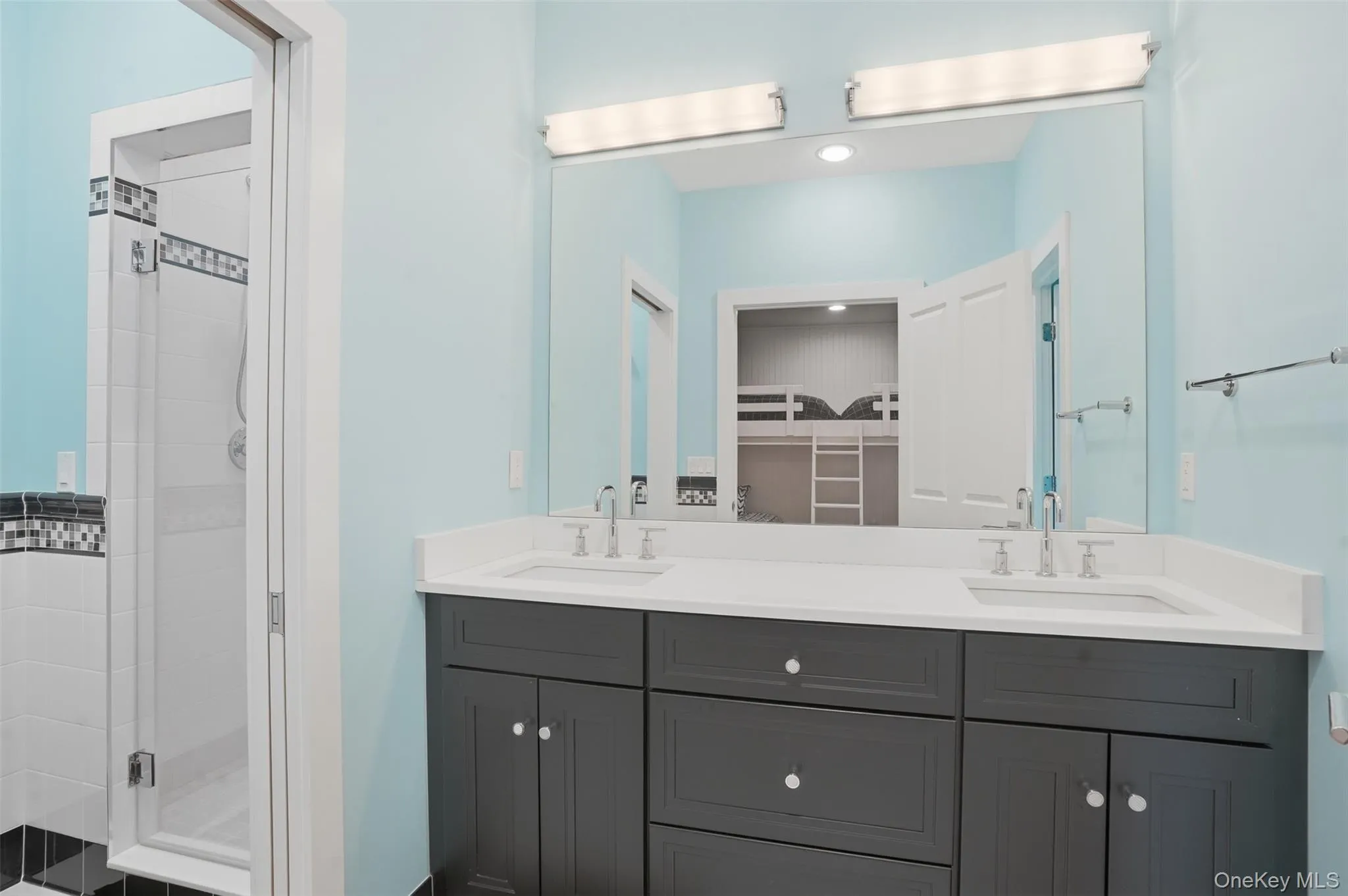Bathroom featuring a double vanity with dark cabinetry and a light-toned countertop, dual sinks with chrome fixtures, and a large wall-mounted mirror Bathroom featuring a double vanity with dark cabinetry and a light-toned countertop, dual sinks with chrome fixtures, and a large wall-mounted mirror
