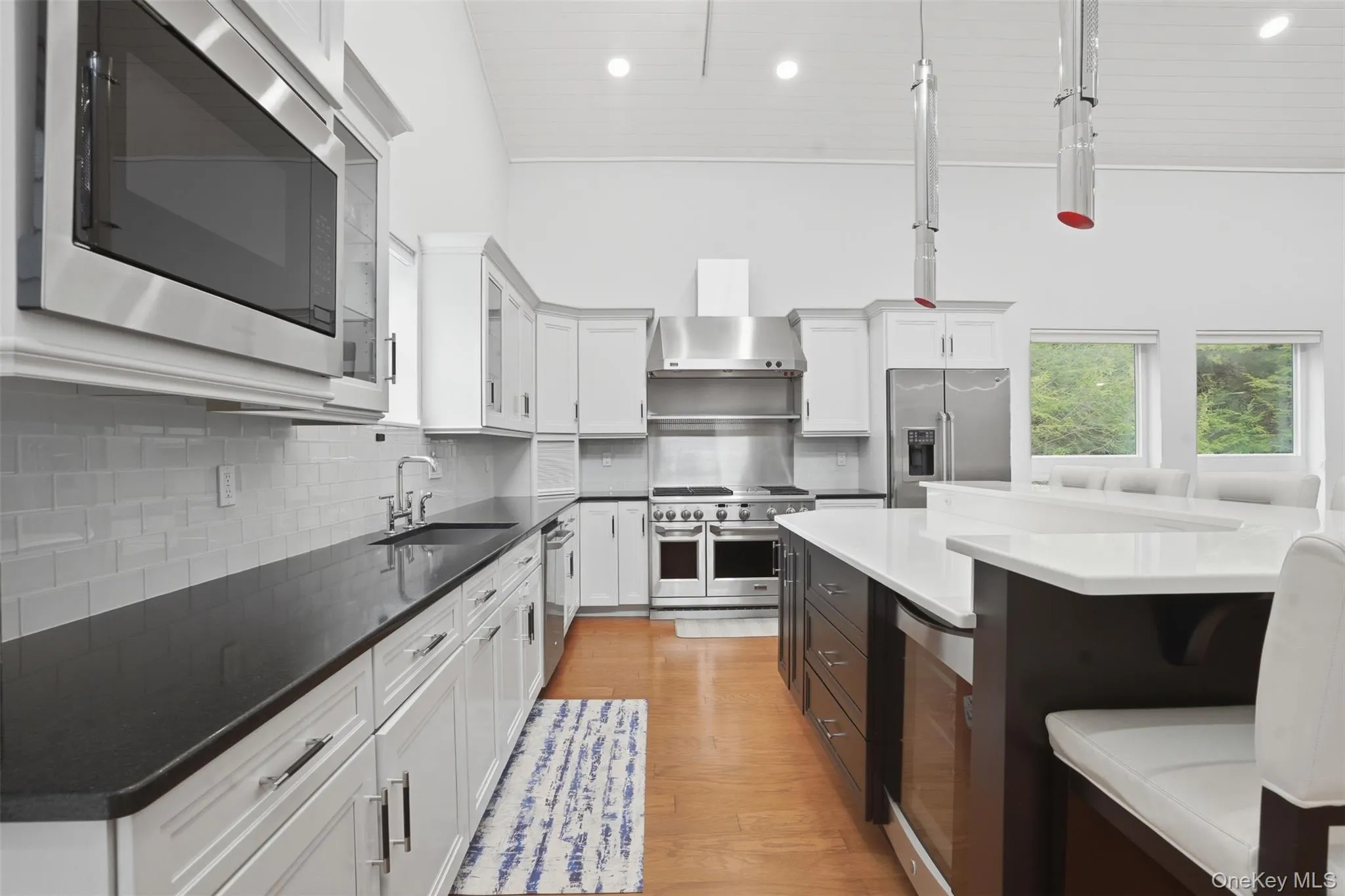 High-ceiling kitchen featuring white cabinetry, stainless steel appliances, dark countertops, and a central island with integrated seating High-ceiling kitchen featuring white cabinetry, stainless steel appliances, dark countertops, and a central island with integrated seating