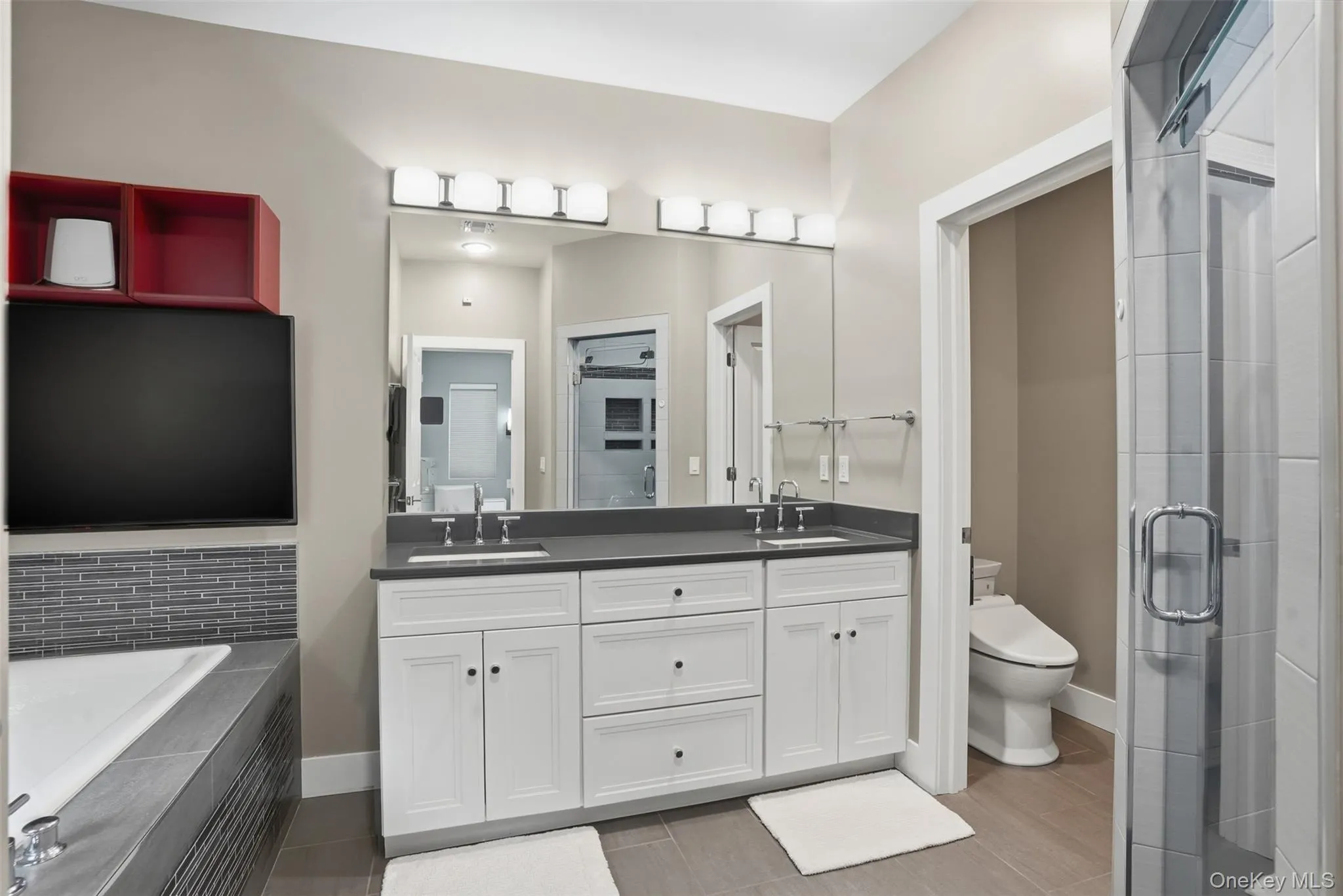 Bathroom featuring a dual-sink vanity with white cabinetry and dark countertop, large wall-mounted mirror, recessed bathtub with gray tile surround and mosaic accent, and a glass-enclosed shower Bathroom featuring a dual-sink vanity with white cabinetry and dark countertop, large wall-mounted mirror, recessed bathtub with gray tile surround and mosaic accent, and a glass-enclosed shower