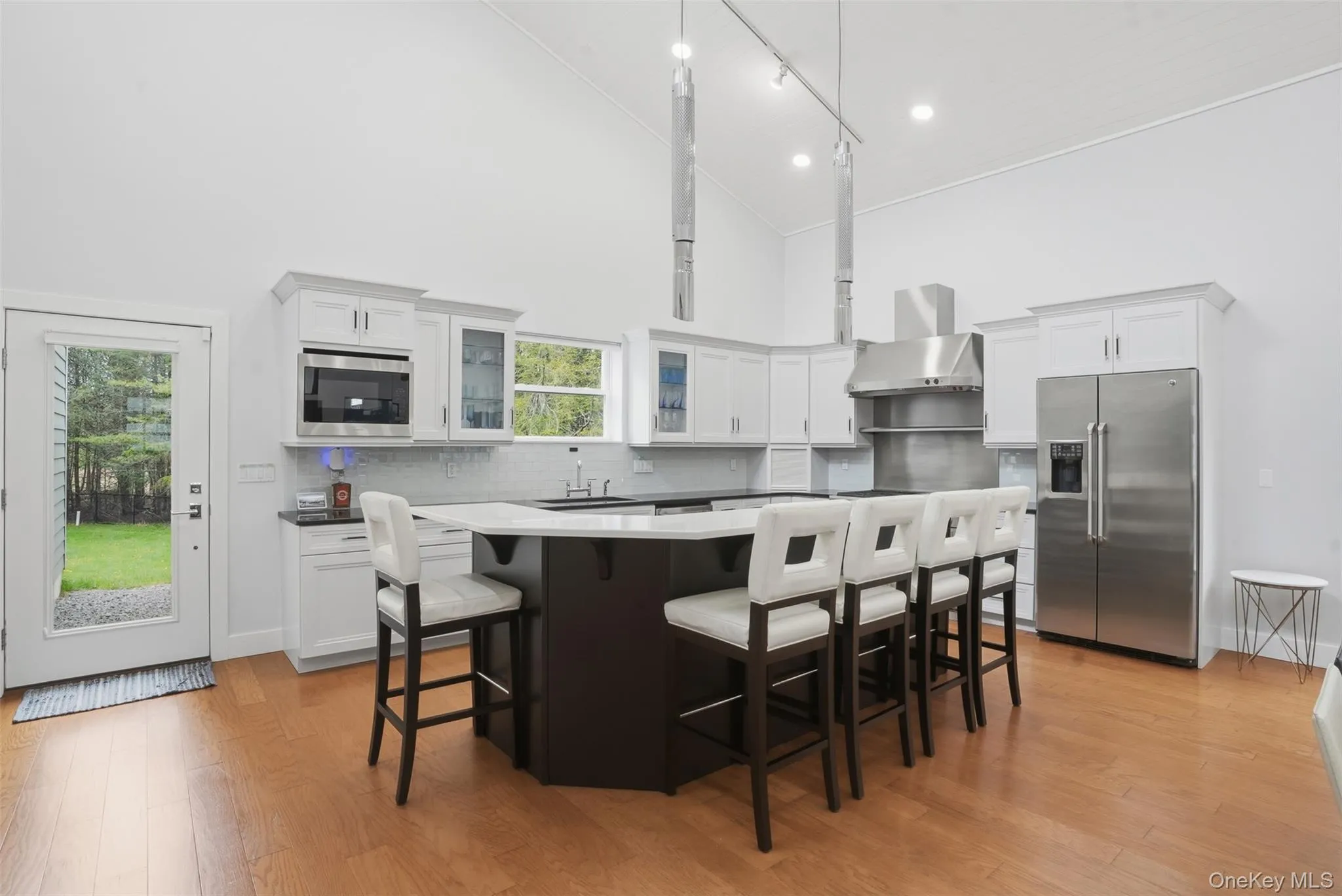 Vaulted ceiling kitchen featuring an expansive central island with seating, stainless steel appliances, white cabinetry, and wood-finish flooring Vaulted ceiling kitchen featuring an expansive central island with seating, stainless steel appliances, white cabinetry, and wood-finish flooring