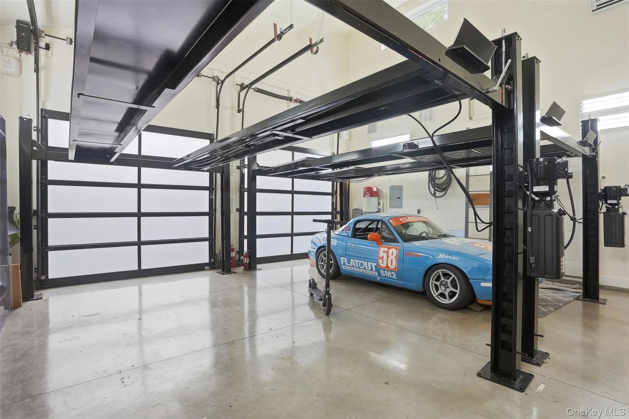 Spacious garage featuring light concrete flooring, multiple overhead garage doors with frosted panels, and two black metal car lifts Spacious garage featuring light concrete flooring, multiple overhead garage doors with frosted panels, and two black metal car lifts