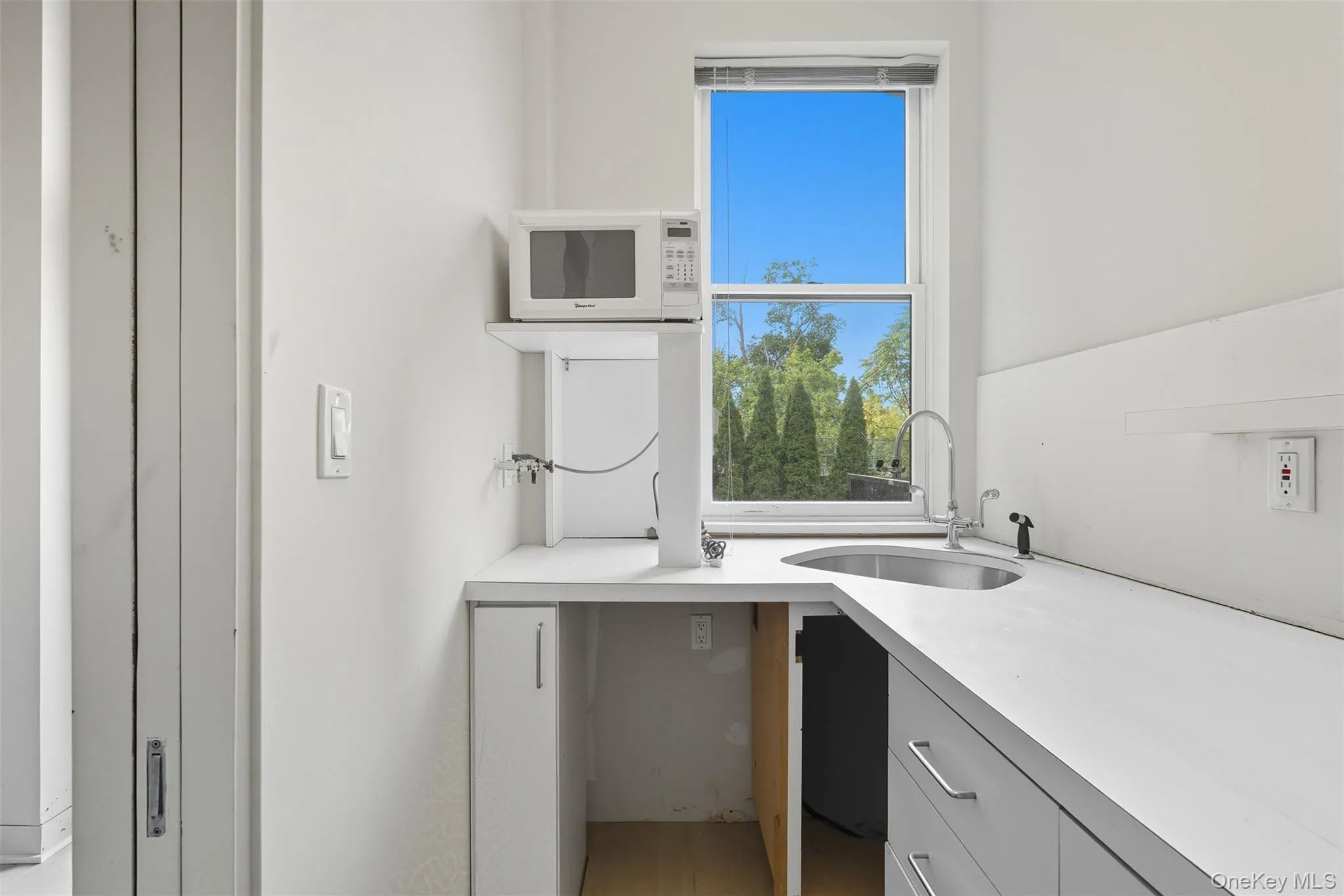 Compact kitchenette featuring white cabinetry, a stainless steel sink with a gooseneck faucet, and a window with outdoor tree scenery Compact kitchenette featuring white cabinetry, a stainless steel sink with a gooseneck faucet, and a window with outdoor tree scenery