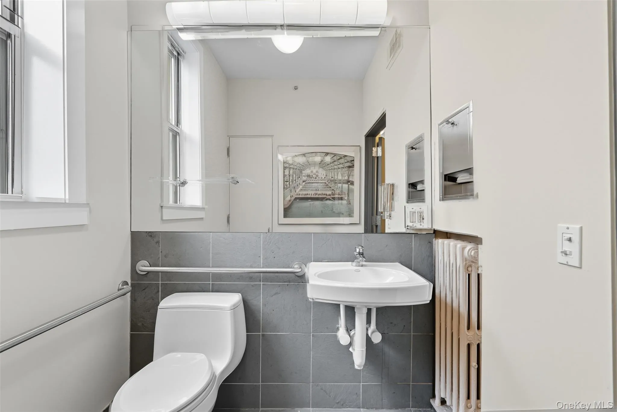 Bathroom featuring a wall-mounted sink, a large wall mirror, gray tile wainscoting, and a window for natural light Bathroom featuring a wall-mounted sink, a large wall mirror, gray tile wainscoting, and a window for natural light
