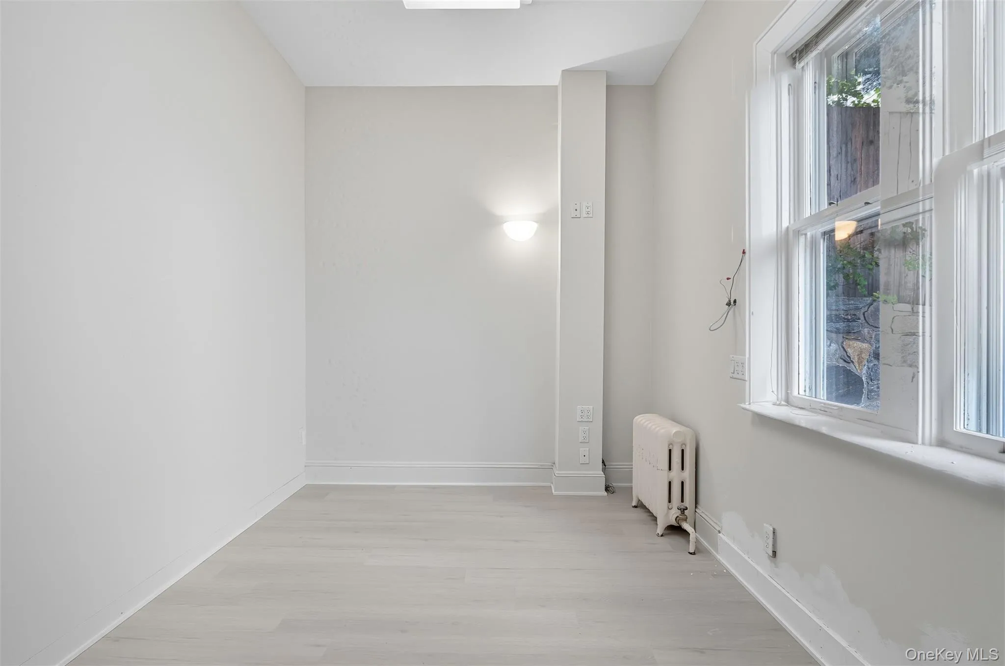 Room featuring two windows, light wood-finish flooring, a wall-mounted light fixture, white baseboards, and a radiator Room featuring two windows, light wood-finish flooring, a wall-mounted light fixture, white baseboards, and a radiator