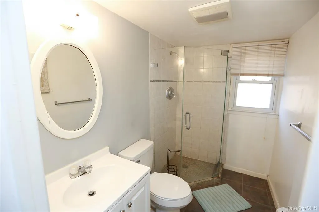 Bathroom featuring a glass-enclosed shower, white vanity with integrated sink, oval mirror, wall-mounted light fixture, and a window with blinds Bathroom featuring a glass-enclosed shower, white vanity with integrated sink, oval mirror, wall-mounted light fixture, and a window with blinds