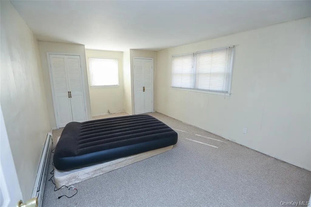 Carpeted room featuring two windows with blinds, two louvered bifold closet doors, and wall-mounted baseboard heating Carpeted room featuring two windows with blinds, two louvered bifold closet doors, and wall-mounted baseboard heating