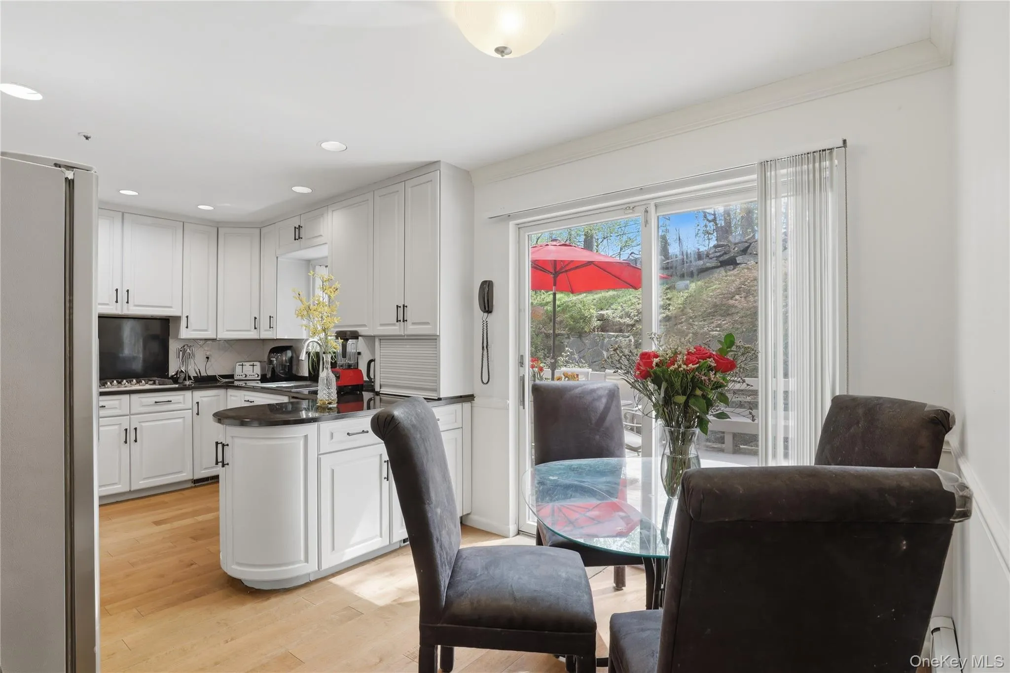 Bright kitchen featuring white cabinetry, dark countertops, a central island, wood-finish flooring, and sliding glass doors opening to the exterior Bright kitchen featuring white cabinetry, dark countertops, a central island, wood-finish flooring, and sliding glass doors opening to the exterior