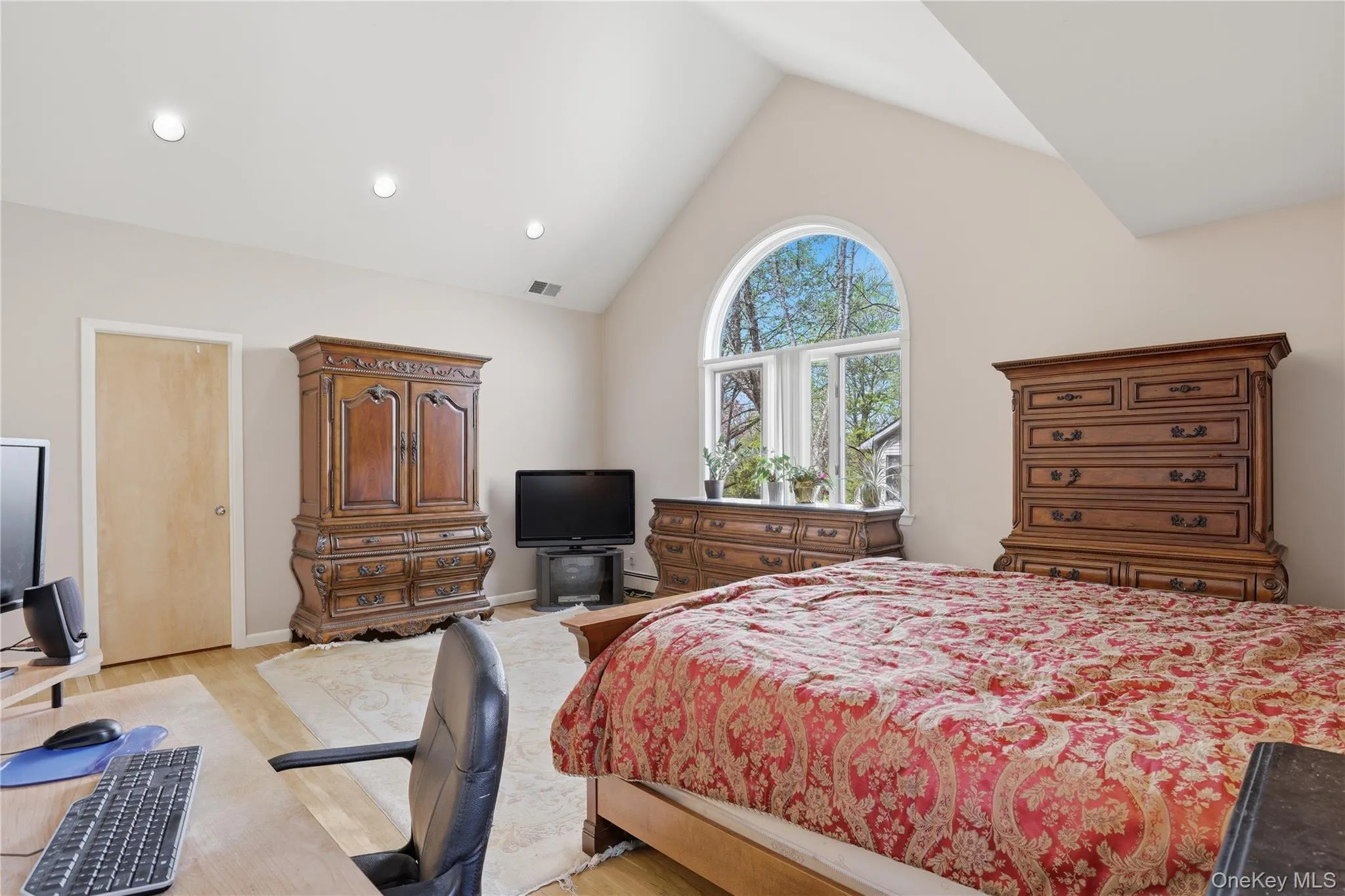 Vaulted ceiling primary bedroom featuring a large arched window, multiple recessed light fixtures, light wood-finish flooring, and a neutral wall color Vaulted ceiling primary bedroom featuring a large arched window, multiple recessed light fixtures, light wood-finish flooring, and a neutral wall color
