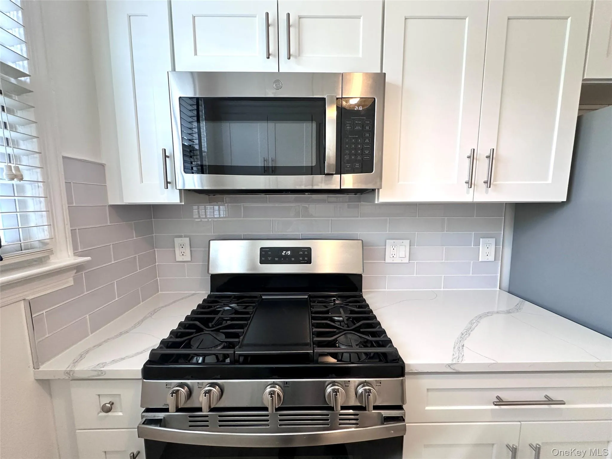 Modern kitchen featuring white shaker-style cabinetry, stainless steel appliances including an oven range and microwave, light-toned countertops with veining, and a grey subway tile backsplash Modern kitchen featuring white shaker-style cabinetry, stainless steel appliances including an oven range and microwave, light-toned countertops with veining, and a grey subway tile backsplash