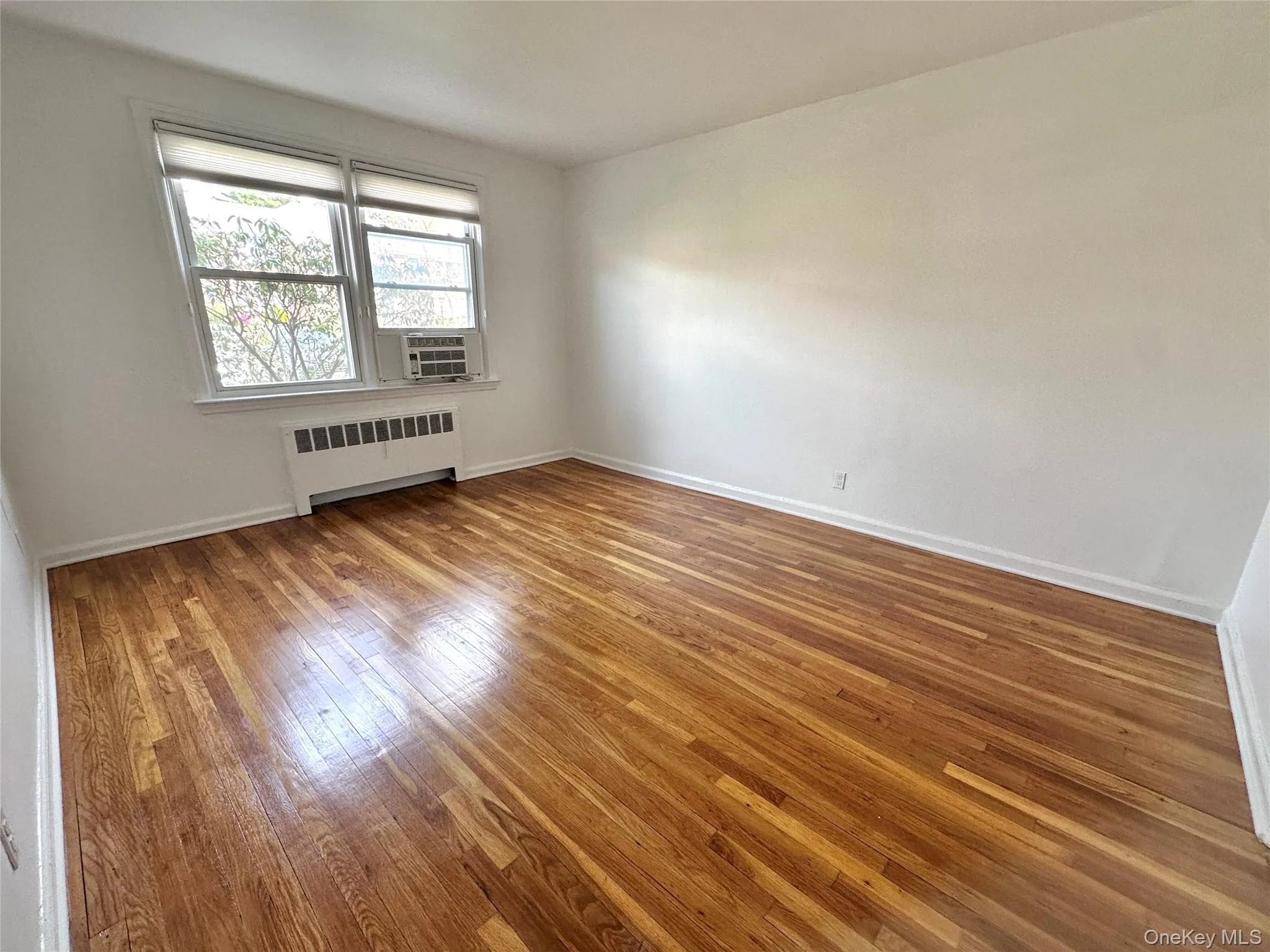 Bright room featuring wood flooring, white walls, and a window with roller shades; AC included Bright room featuring wood flooring, white walls, and a window with roller shades; AC included