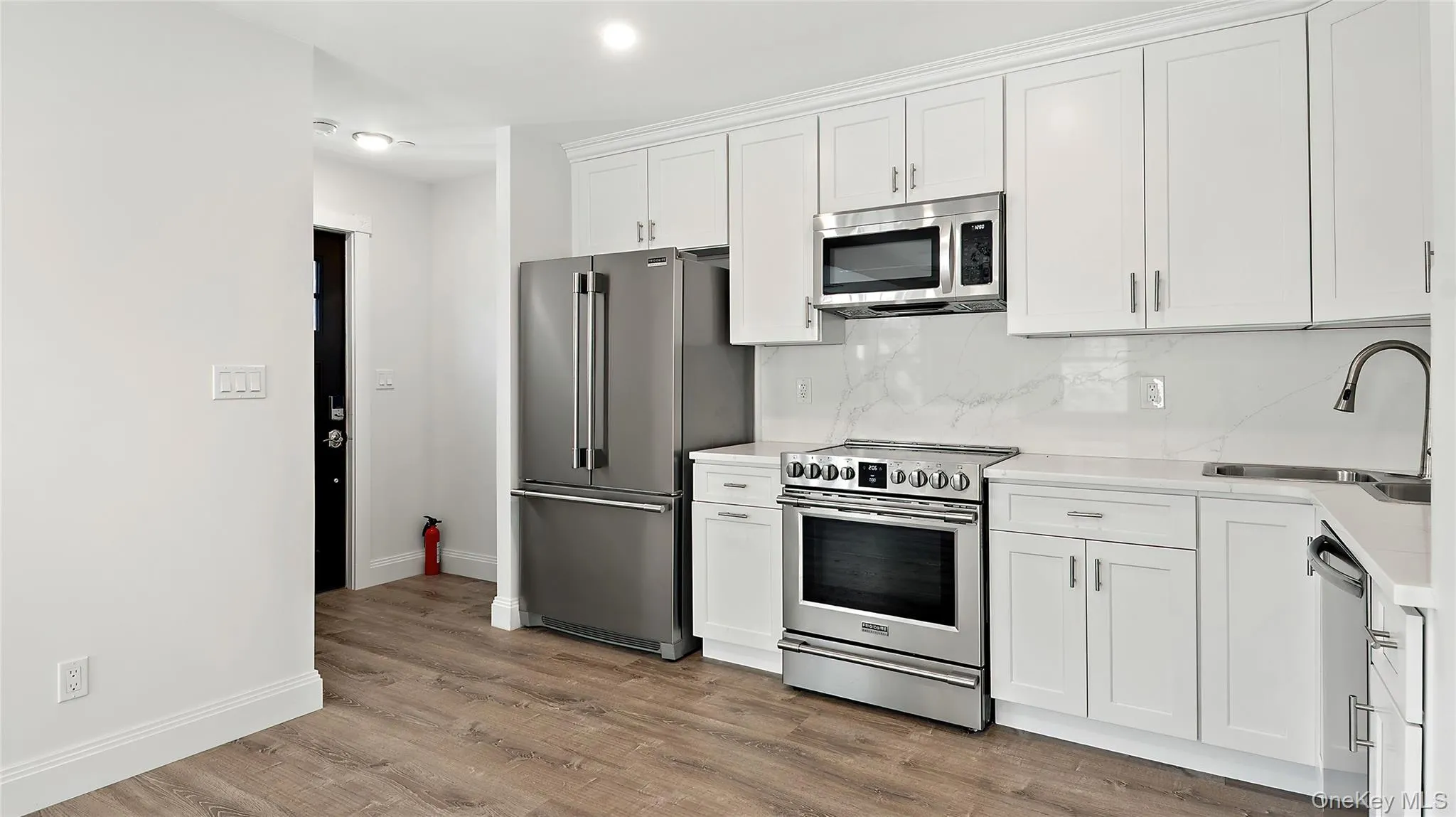 Modern kitchen featuring white shaker cabinetry, stainless steel appliances, white countertops, a tiled backsplash, and wood-finish flooring Modern kitchen featuring white shaker cabinetry, stainless steel appliances, white countertops, a tiled backsplash, and wood-finish flooring