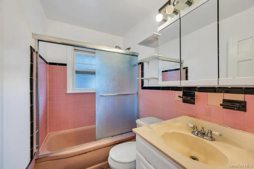 Bathroom featuring a built-in bathtub with pink and black tiled surround, a sliding glass shower door, and a single window Bathroom featuring a built-in bathtub with pink and black tiled surround, a sliding glass shower door, and a single window