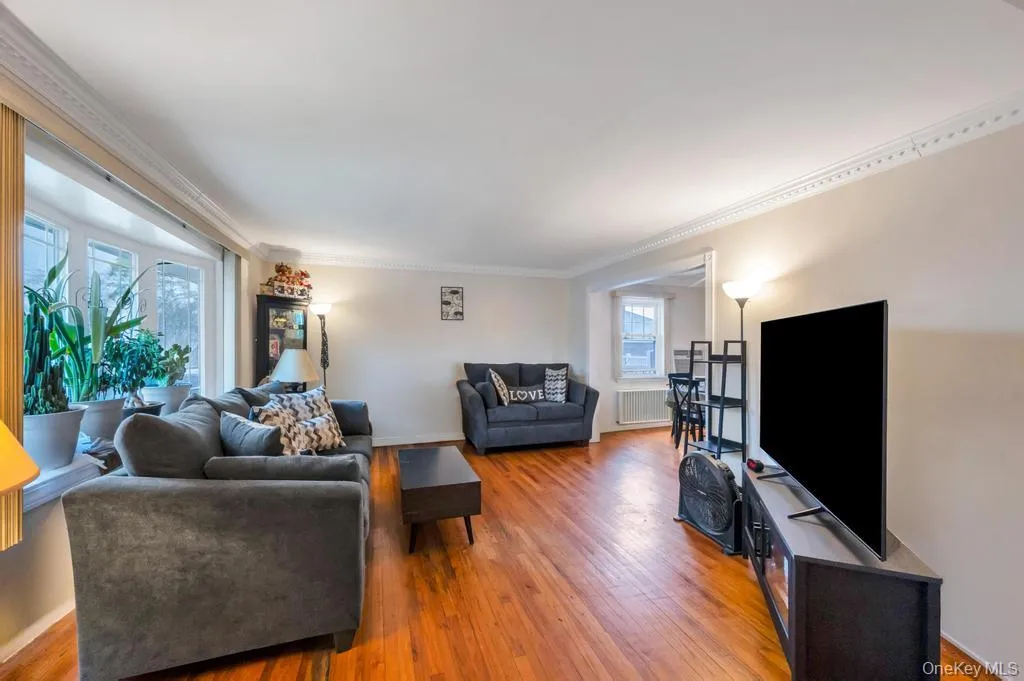Expansive living area featuring warm wood-finish flooring, a large bay window, and detailed crown molding Expansive living area featuring warm wood-finish flooring, a large bay window, and detailed crown molding
