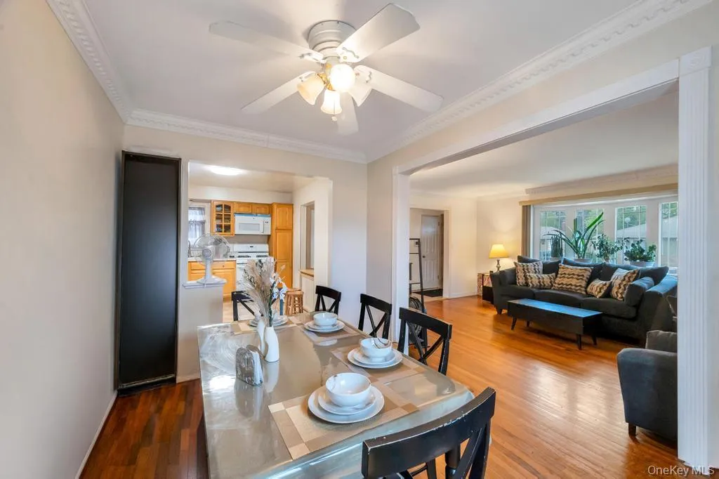 Dining area featuring crown molding, hardwood flooring, and an open pass-through to the kitchen with wood cabinetry and white appliances Dining area featuring crown molding, hardwood flooring, and an open pass-through to the kitchen with wood cabinetry and white appliances