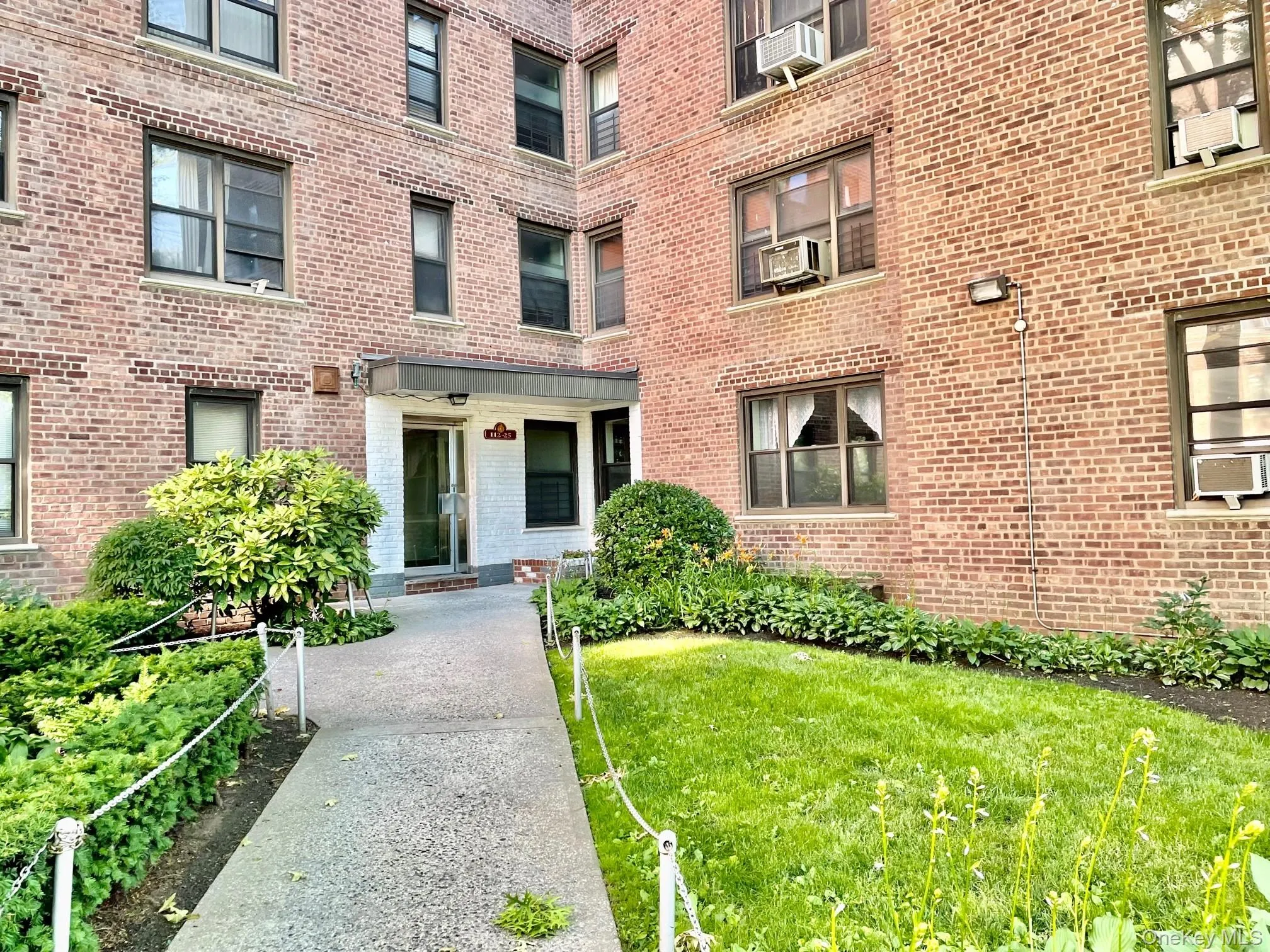 Brick facade building featuring a central entry, a concrete walkway, mature shrubbery, and a manicured lawn Brick facade building featuring a central entry, a concrete walkway, mature shrubbery, and a manicured lawn