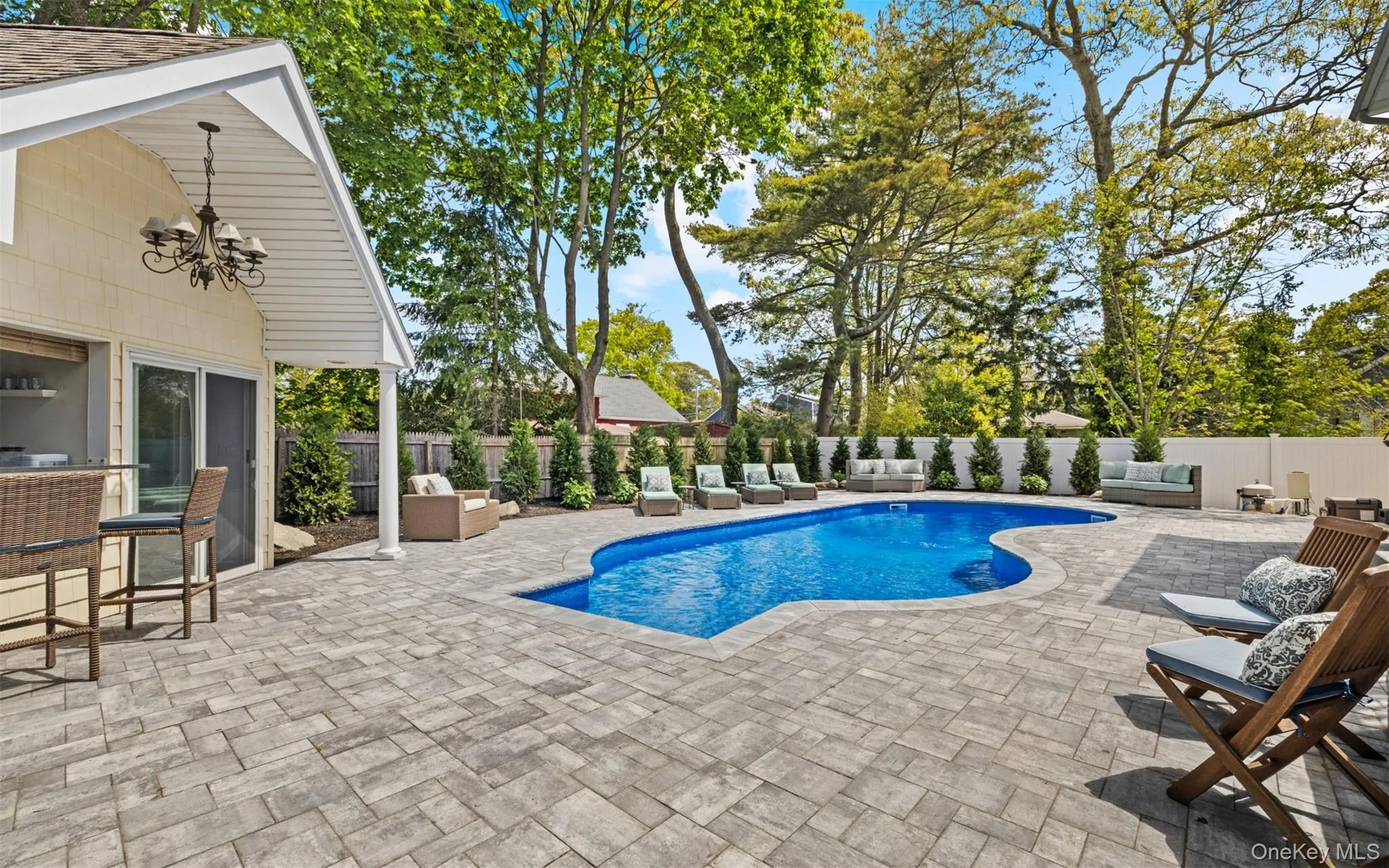 Backyard oasis featuring a freeform in-ground pool, extensive paver patio, mature landscaping, and a covered outdoor bar area with pendant lighting Backyard oasis featuring a freeform in-ground pool, extensive paver patio, mature landscaping, and a covered outdoor bar area with pendant lighting