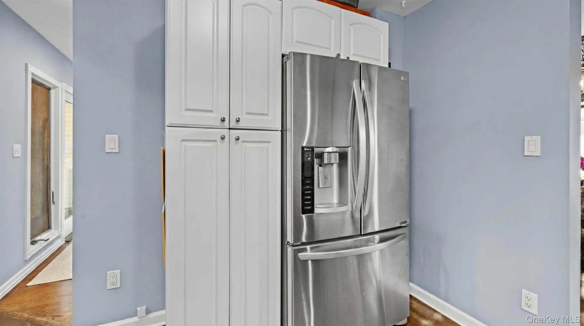 Modern kitchen area featuring a stainless steel French door refrigerator, white shaker-style cabinetry, wood-finish flooring, and light blue walls Modern kitchen area featuring a stainless steel French door refrigerator, white shaker-style cabinetry, wood-finish flooring, and light blue walls