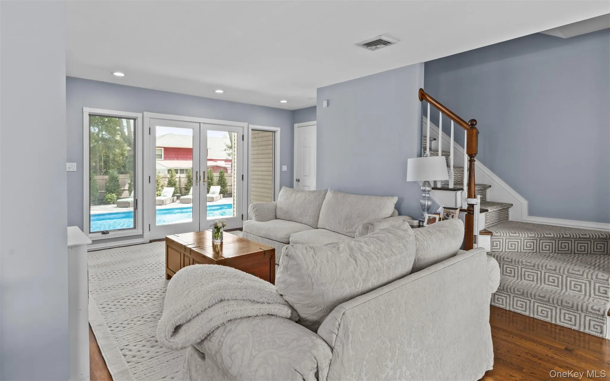 Light-filled living room with French doors opening to an outdoor pool area, recessed lighting, wood-flooring, and a carpeted staircase with a wood handrail Light-filled living room with French doors opening to an outdoor pool area, recessed lighting, wood-flooring, and a carpeted staircase with a wood handrail