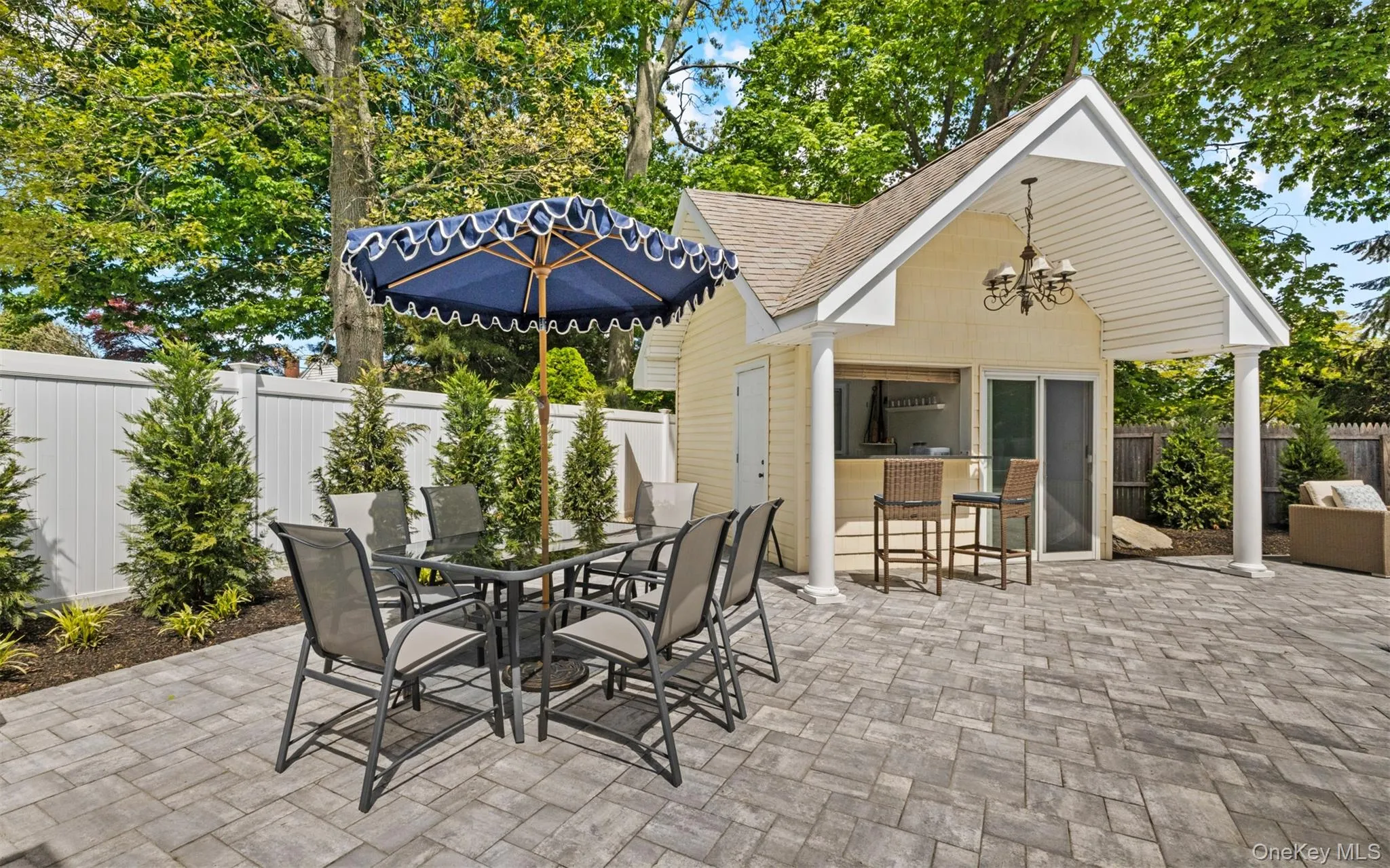Expansive paver patio with a dedicated pool house featuring an outdoor bar area, covered porch, glass sliding door,and chandelier lighting Expansive paver patio with a dedicated pool house featuring an outdoor bar area, covered porch, glass sliding door,and chandelier lighting