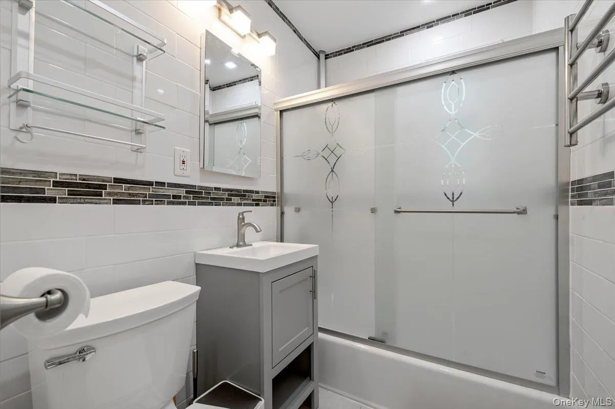 Bathroom featuring a sliding glass shower door with etched detailing, a single vanity with a gray cabinet, a chrome faucet, and a wall-mounted mirror with integrated lighting Bathroom featuring a sliding glass shower door with etched detailing, a single vanity with a gray cabinet, a chrome faucet, and a wall-mounted mirror with integrated lighting