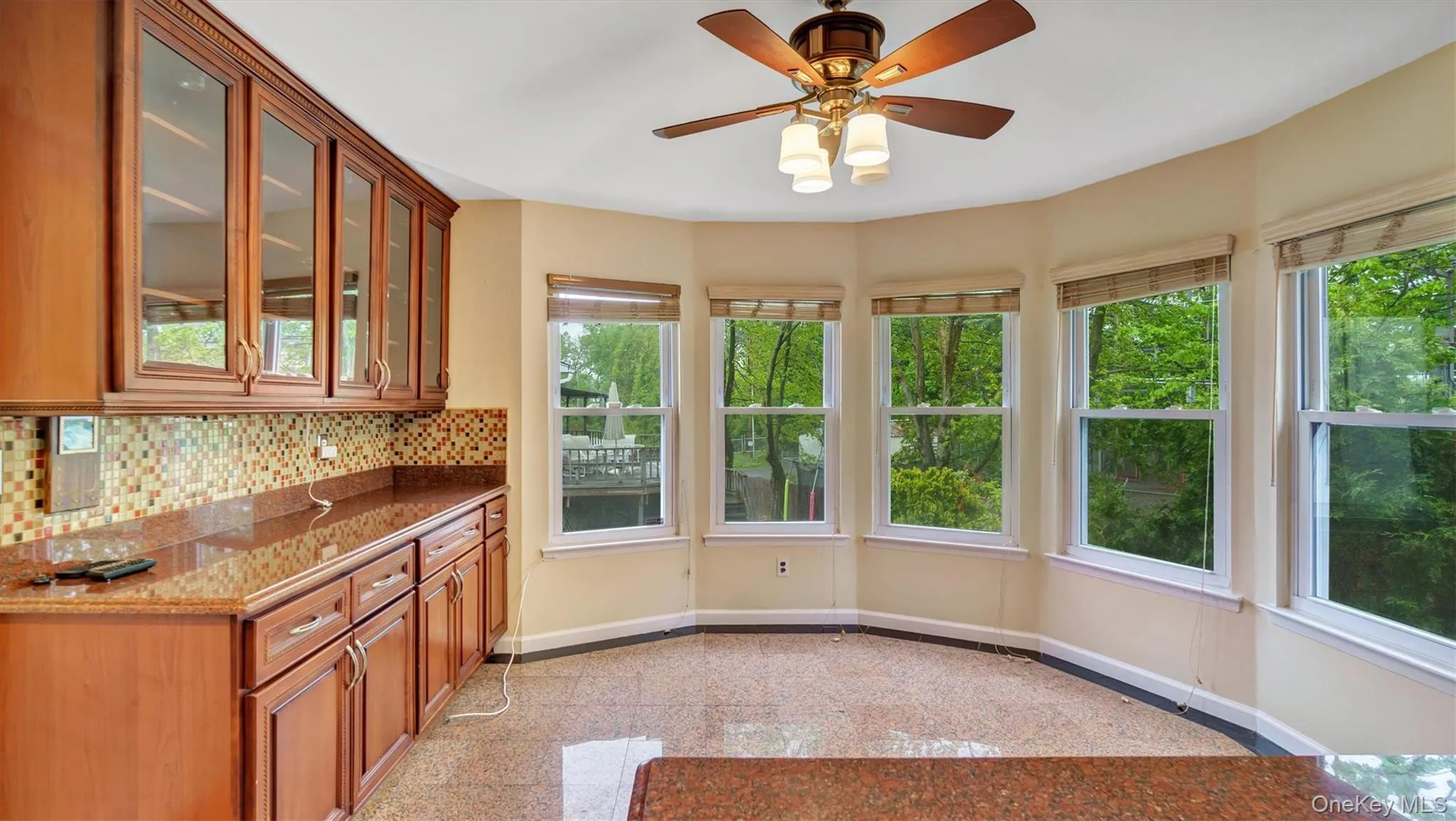 Kitchen featuring brown cabinetry, backsplash, granite finish floor, glass insert cabinets, and baseboards Kitchen featuring brown cabinetry, backsplash, granite finish floor, glass insert cabinets, and baseboards