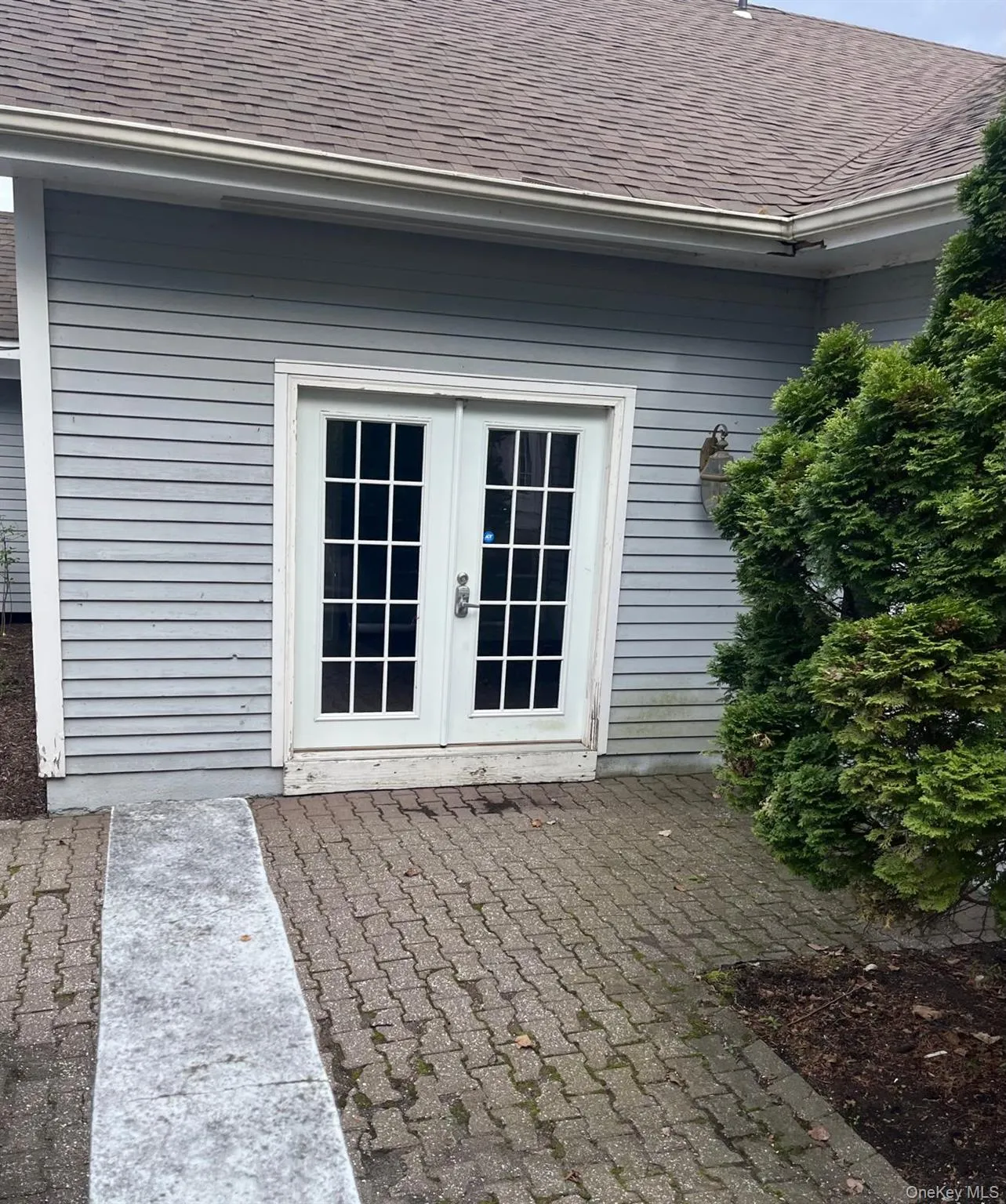 Double French doors with divided lites, light gray horizontal siding, paver patio, brown shingle roof, and a mature evergreen shrub Double French doors with divided lites, light gray horizontal siding, paver patio, brown shingle roof, and a mature evergreen shrub