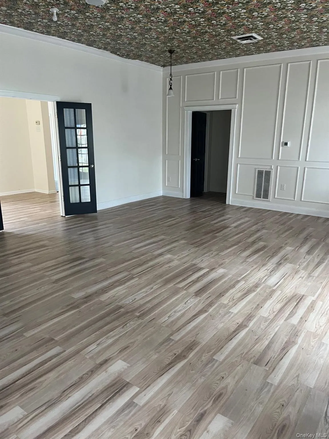 Spacious room featuring wood-finish flooring, a decorative patterned ceiling, and white wall paneling Spacious room featuring wood-finish flooring, a decorative patterned ceiling, and white wall paneling