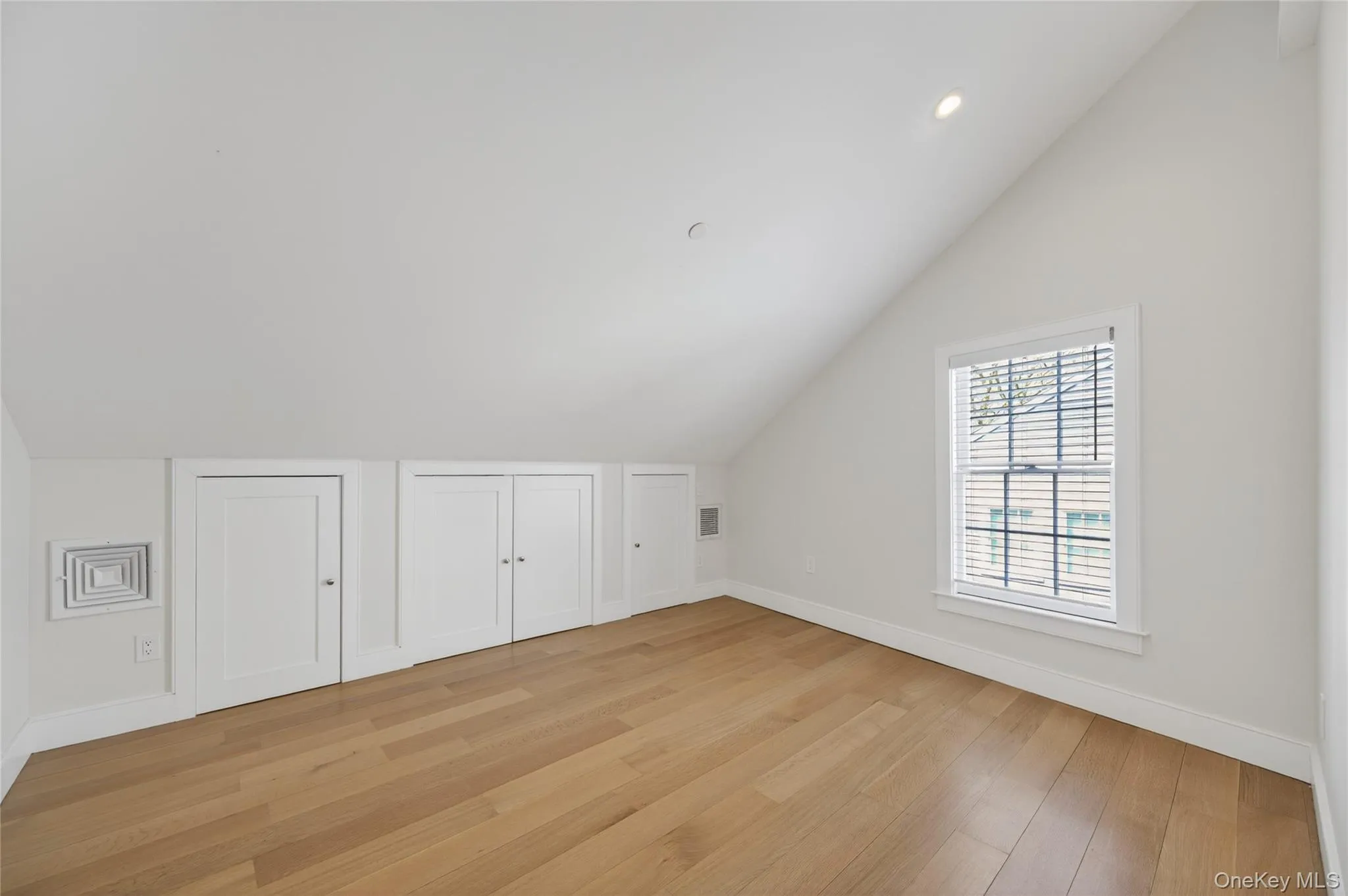 Bright space featuring wood-finish flooring, an angled ceiling, built-in storage doors, a window with blinds, and a recessed ceiling light Bright space featuring wood-finish flooring, an angled ceiling, built-in storage doors, a window with blinds, and a recessed ceiling light