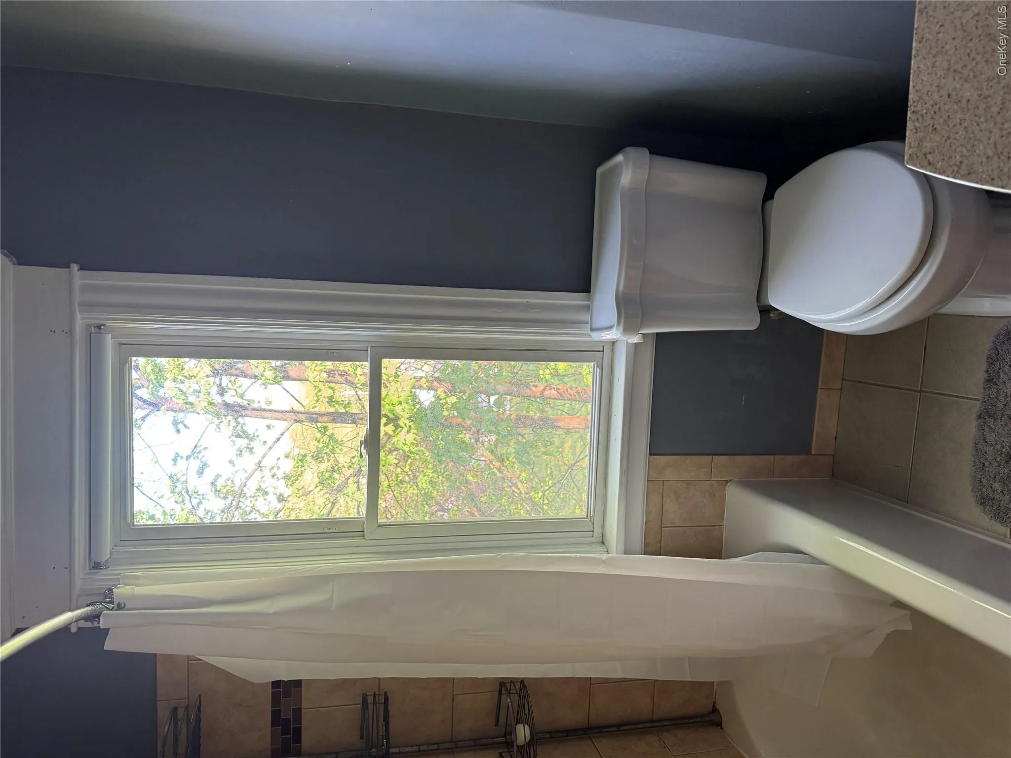 Bathroom featuring a tub/shower combination with ceramic tile surround, a single-pane window with white trim, a standard toilet, and tile flooring Bathroom featuring a tub/shower combination with ceramic tile surround, a single-pane window with white trim, a standard toilet, and tile flooring