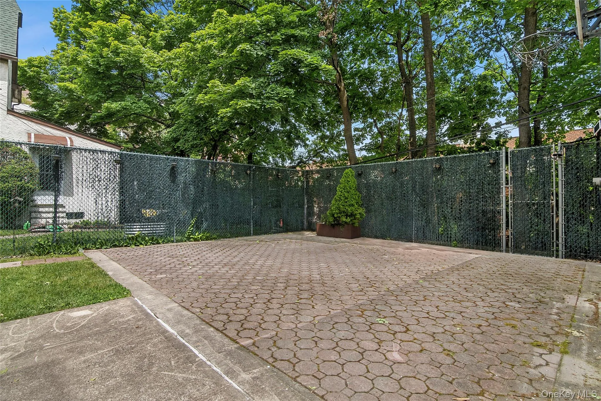 Paved outdoor area featuring hexagonal paver stones, a chain-link fence with privacy screening, and mature trees providing natural canopy Paved outdoor area featuring hexagonal paver stones, a chain-link fence with privacy screening, and mature trees providing natural canopy