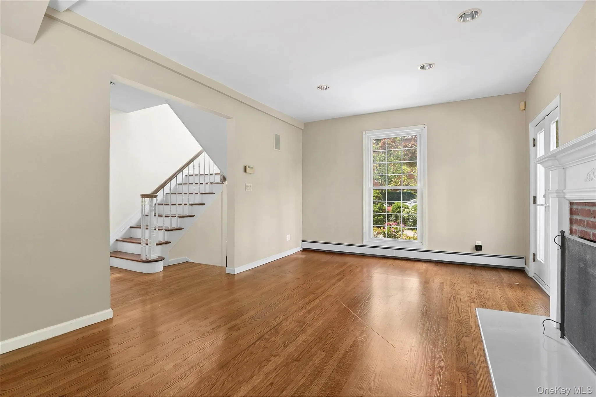 Spacious living area featuring wood-finish flooring, a white mantel fireplace with brick surround, and recessed lighting Spacious living area featuring wood-finish flooring, a white mantel fireplace with brick surround, and recessed lighting