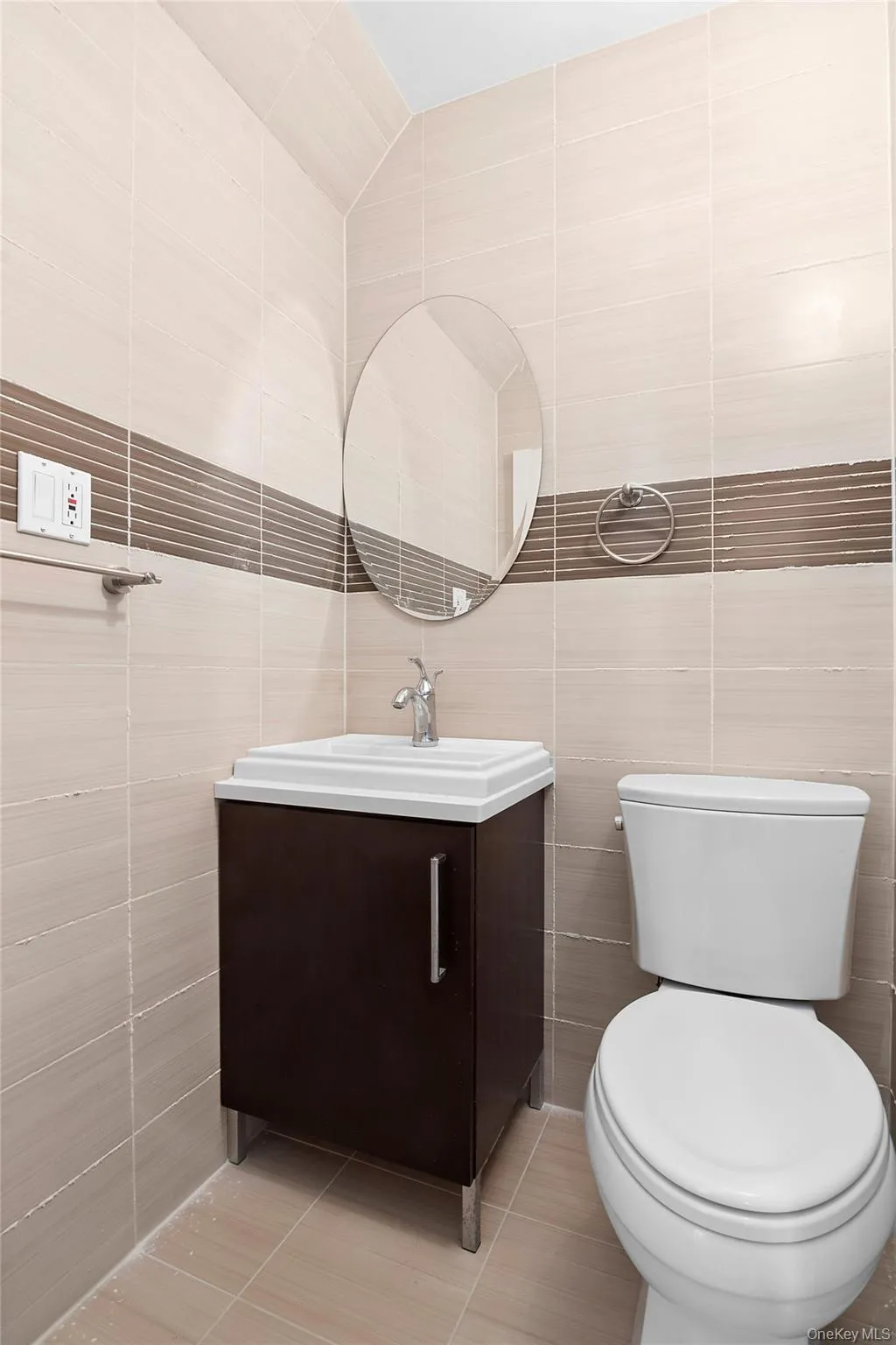 Ensuite Half Bath featuring contemporary light-toned wall tiling with a decorative accent band, a wall-mounted oval mirror, a dark wood-finish vanity with an integrated sink, a chrome faucet, and a standard white toilet Ensuite Half Bath featuring contemporary light-toned wall tiling with a decorative accent band, a wall-mounted oval mirror, a dark wood-finish vanity with an integrated sink, a chrome faucet, and a standard white toilet