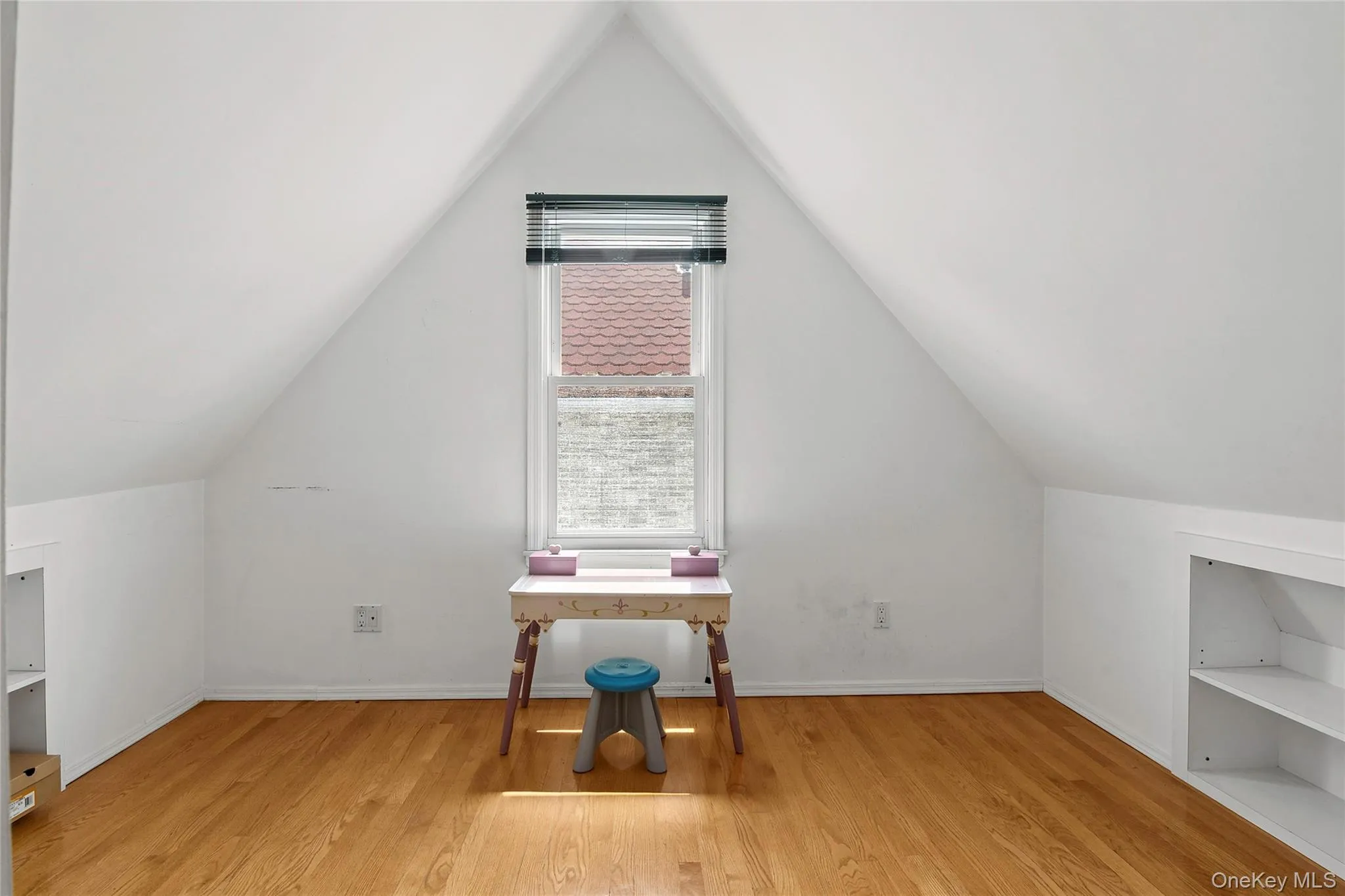 Attic space featuring sloped ceilings, a central window with horizontal blinds, built-in shelving, and wood-finish flooring Attic space featuring sloped ceilings, a central window with horizontal blinds, built-in shelving, and wood-finish flooring