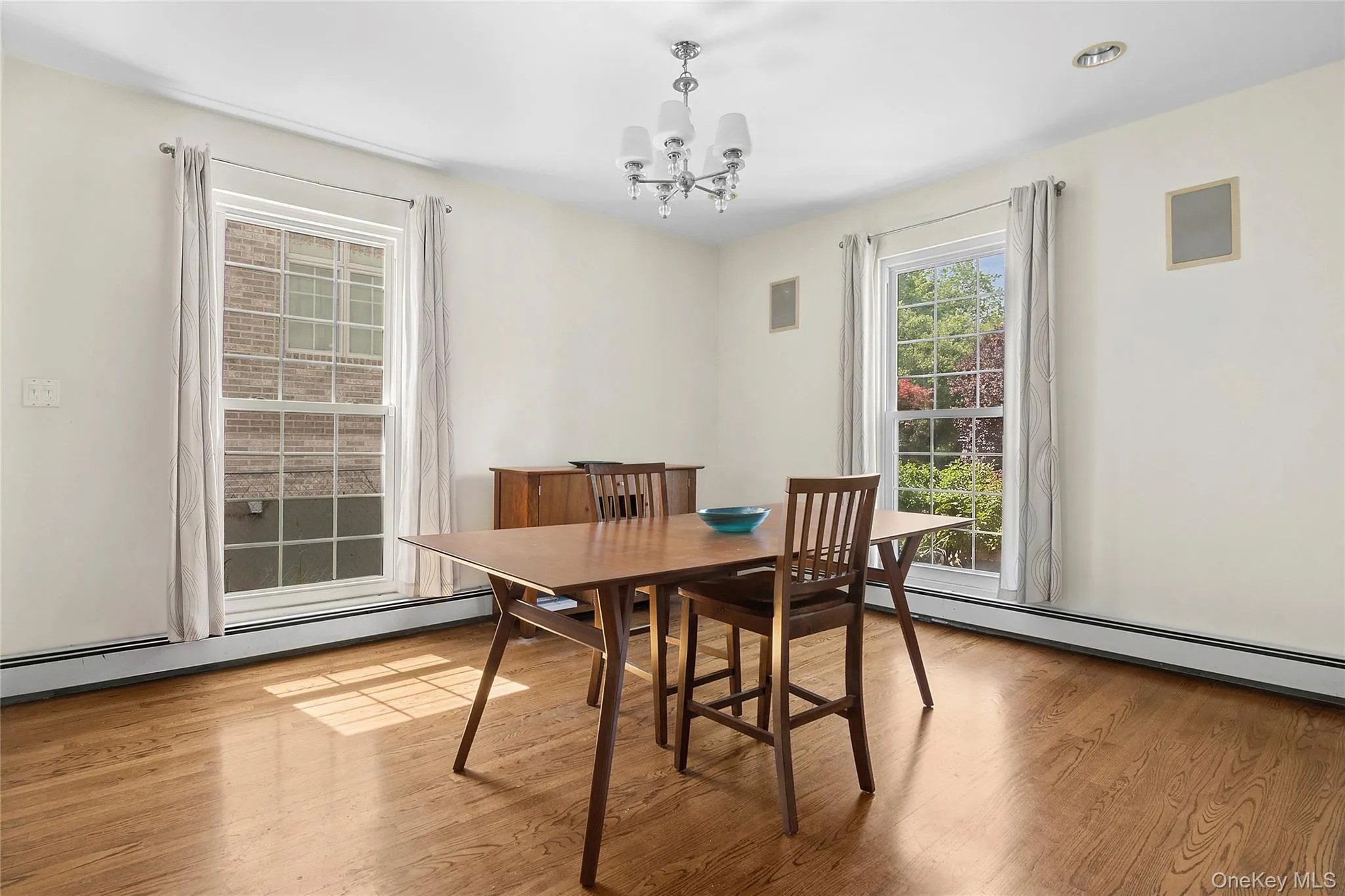 Dining space featuring wood-finish flooring, two large windows, a ceiling-mounted chandelier, and integrated wall speakers Dining space featuring wood-finish flooring, two large windows, a ceiling-mounted chandelier, and integrated wall speakers