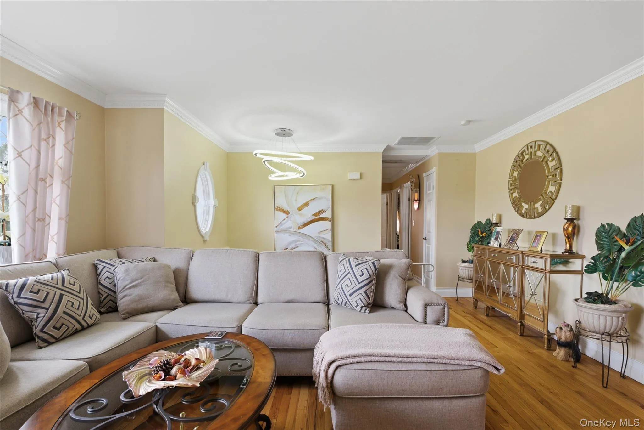 Living area featuring wood-finish flooring, crown molding, a contemporary chandelier, light-toned wall paint, and a window with curtains Living area featuring wood-finish flooring, crown molding, a contemporary chandelier, light-toned wall paint, and a window with curtains