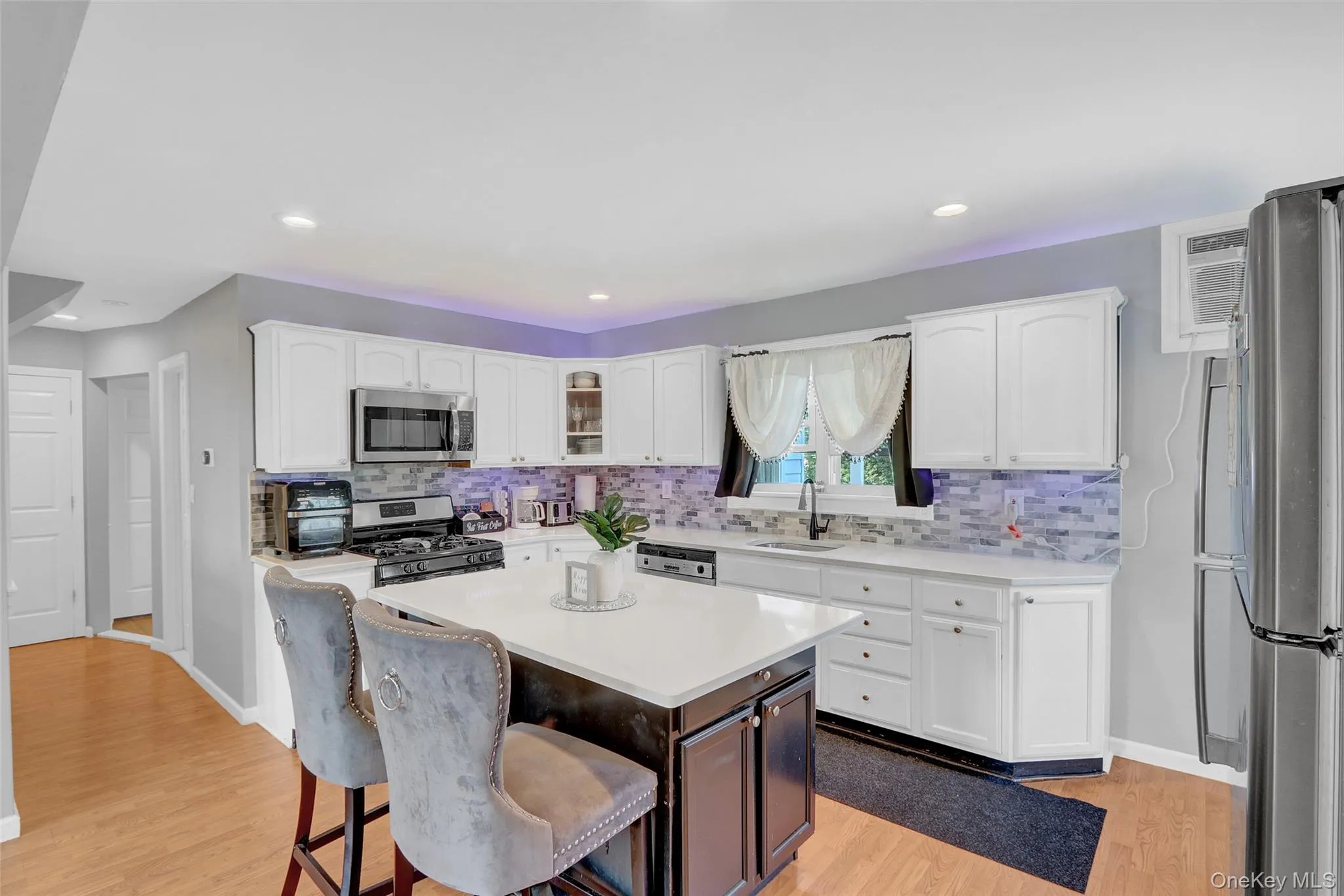 Kitchen featuring white cabinetry with brushed nickel hardware, stainless steel appliances, a white countertop island with seating, a mosaic tile backsplash, and light wood-finish flooring Kitchen featuring white cabinetry with brushed nickel hardware, stainless steel appliances, a white countertop island with seating, a mosaic tile backsplash, and light wood-finish flooring
