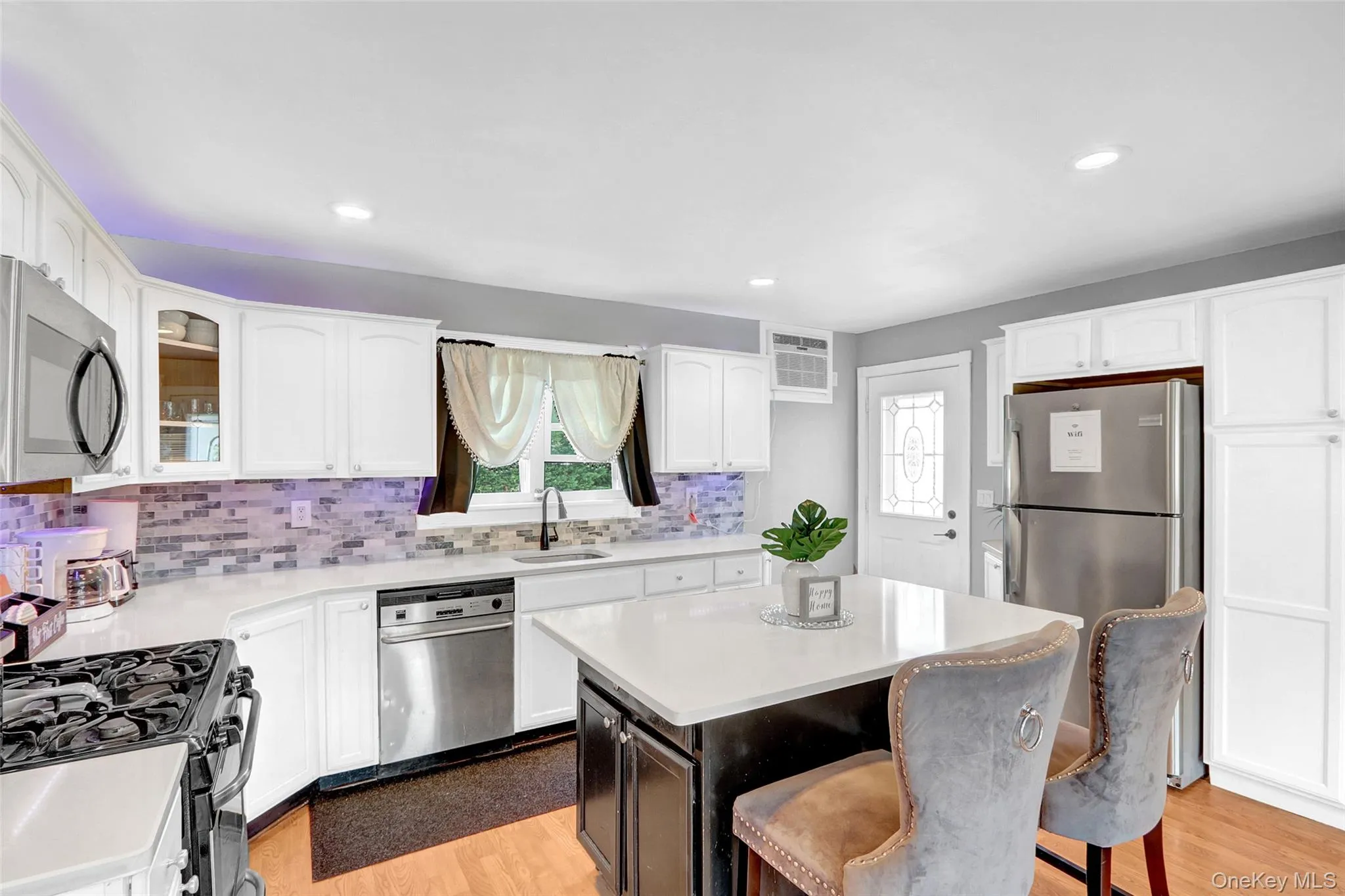 Kitchen featuring white cabinetry, stainless steel appliances, a center island with a light-toned countertop, a multi-tone tile backsplash, and wood-finish flooring Kitchen featuring white cabinetry, stainless steel appliances, a center island with a light-toned countertop, a multi-tone tile backsplash, and wood-finish flooring