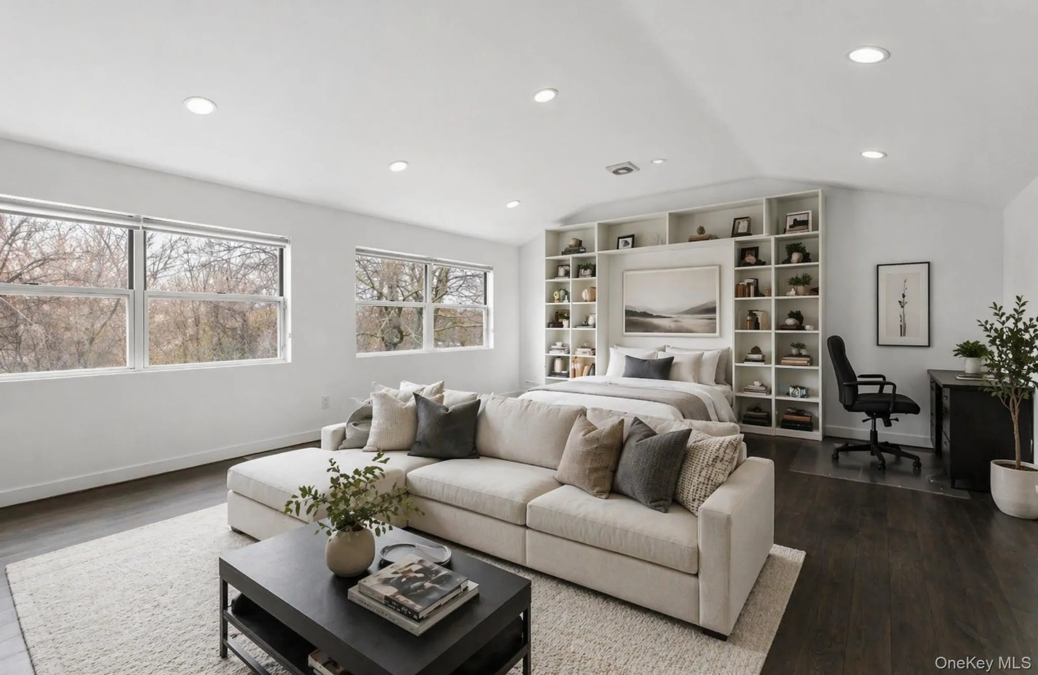 Bright, open-concept living area featuring multiple large windows, dark wood-finish flooring, recessed lighting, and a built-in shelving system with integrated bed frame Bright, open-concept living area featuring multiple large windows, dark wood-finish flooring, recessed lighting, and a built-in shelving system with integrated bed frame